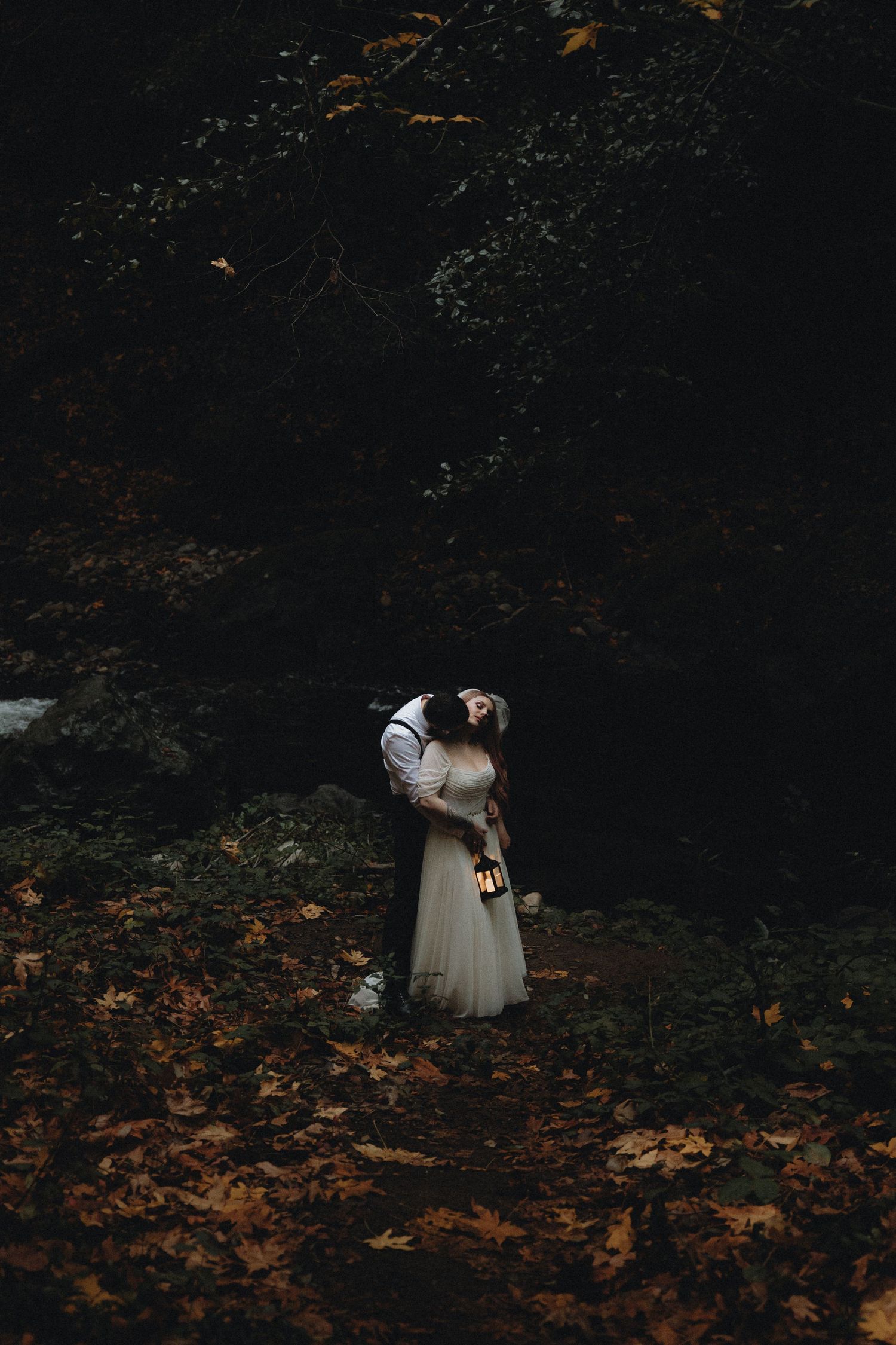 A figure in a white dress walks through a dark moody forest scattered with autumn leaves on the ground.
