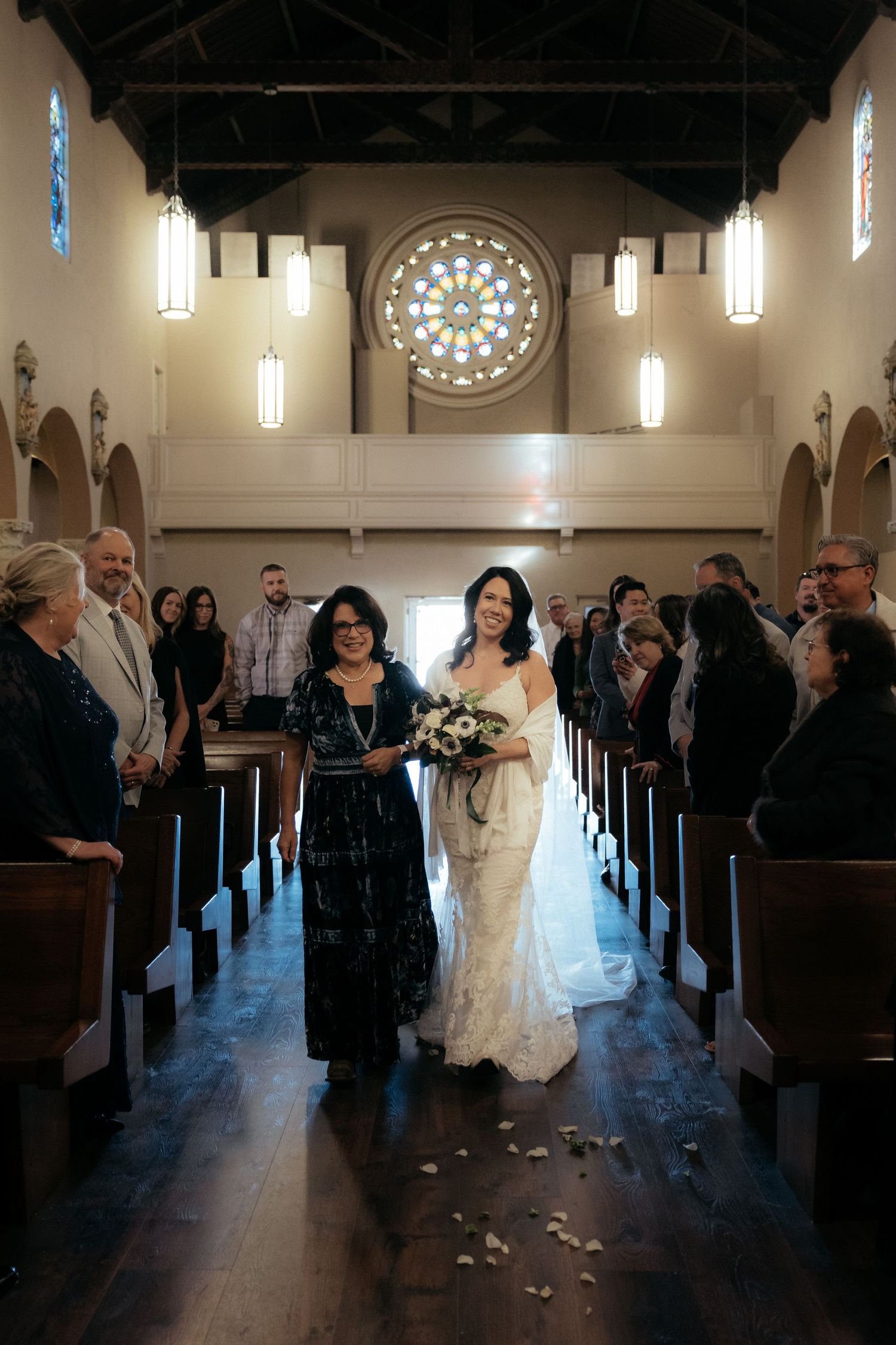 Wedding guests line the aisle of a church sanctuary with hanging pendant lights and a stained glass window.