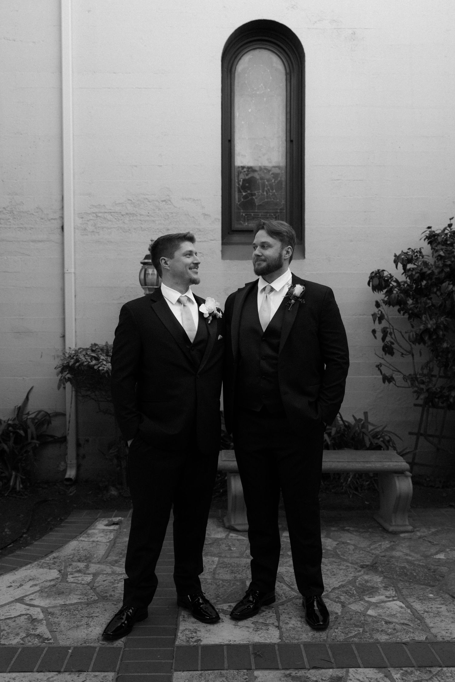Two grooms in black tuxedos share a joyful moment together during their wedding against a white wall with arched window.