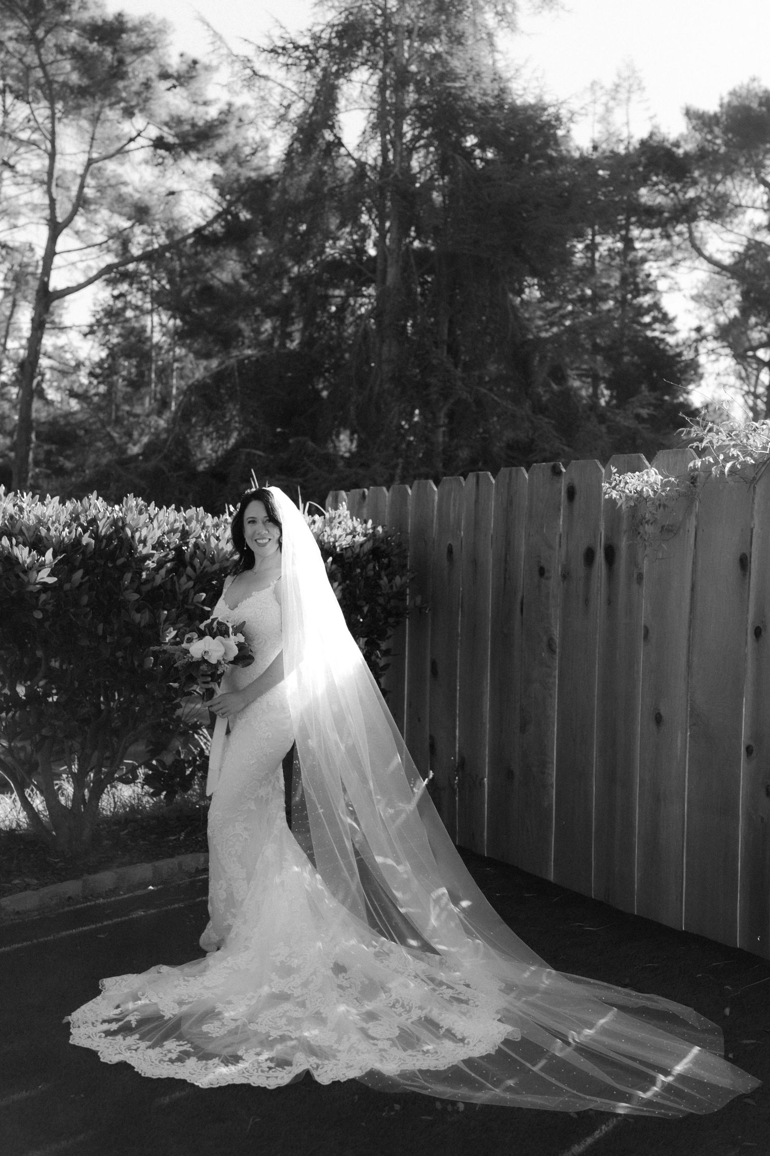 Black and white photo of a bride in a lace wedding dress with long veil standing by a wooden fence.