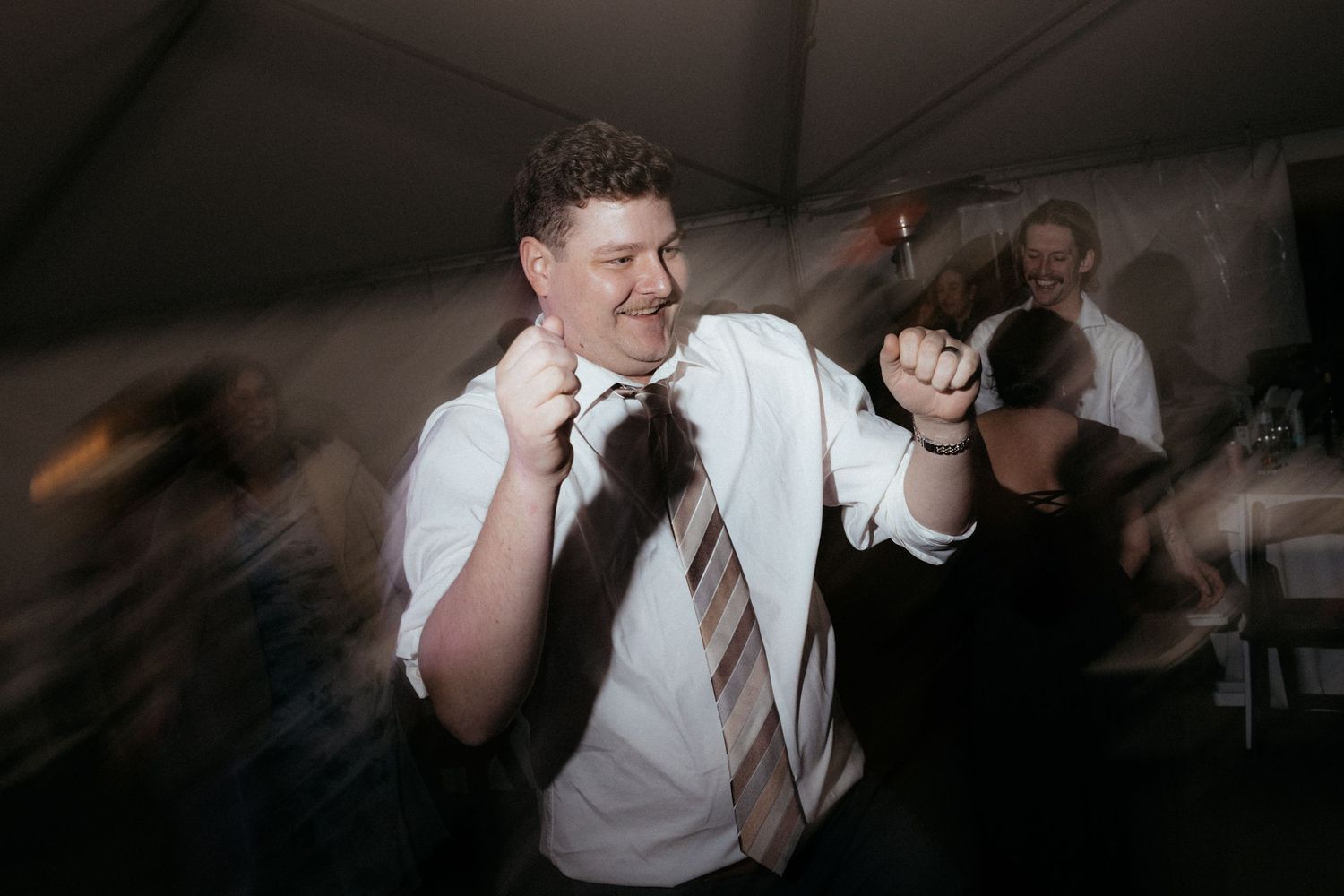 A party guest in white shirt and tie dances enthusiastically under tent lighting.