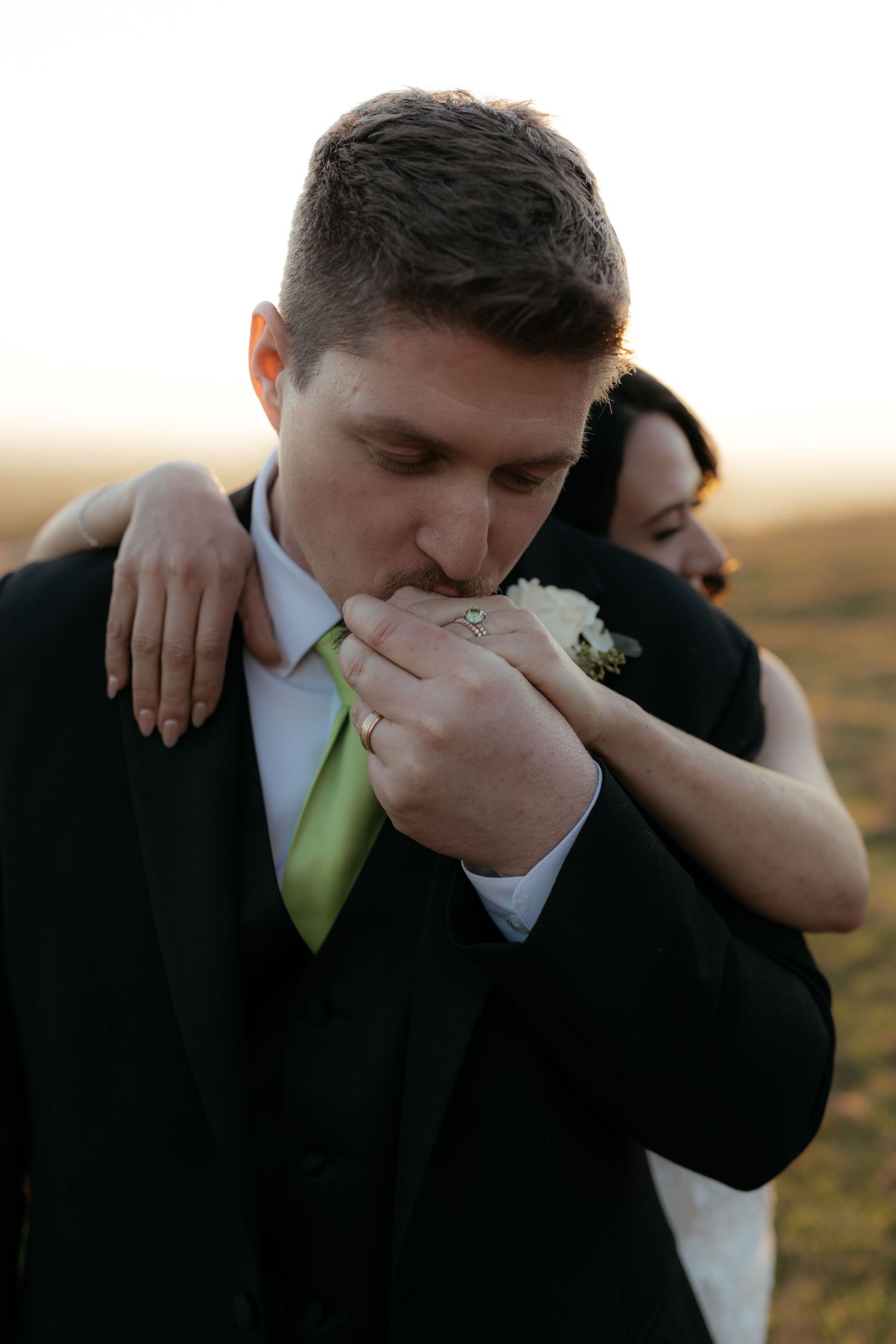 Intimate moment between newlyweds as groom kisses bride's hand during sunset outdoor photo session.