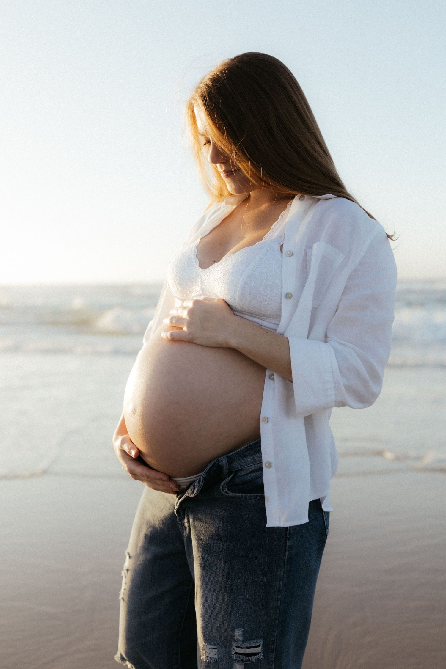 Pregnant woman in white shirt and jeans cradling her baby bump on a sunny beach with ocean waves behind her.