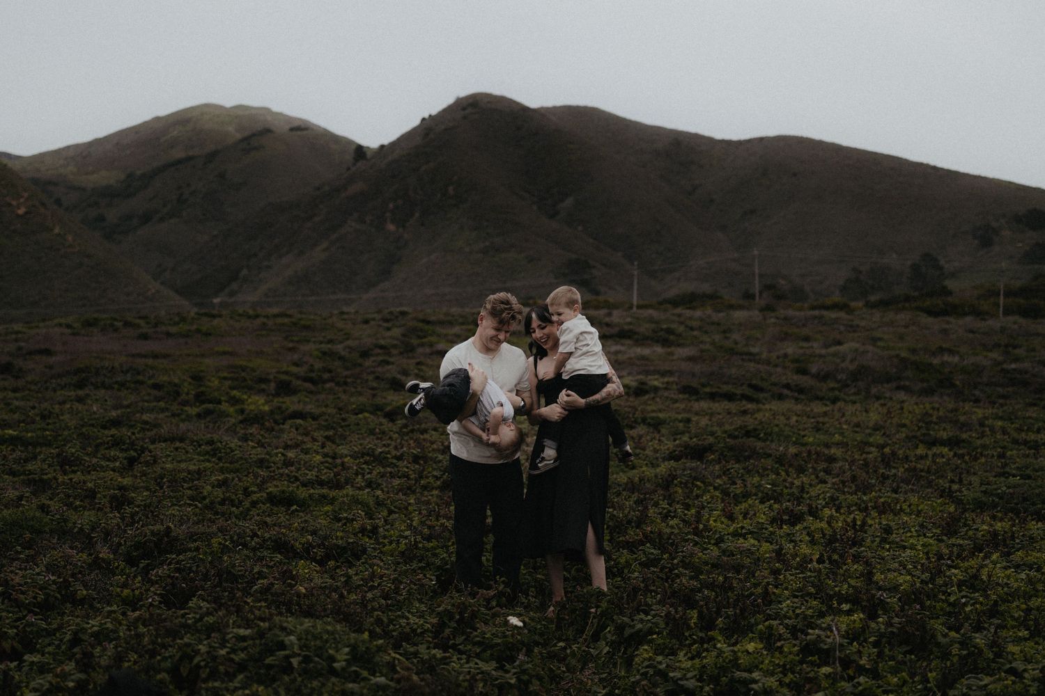 Family of four standing in lush green field with dramatic mountain backdrop during overcast day.