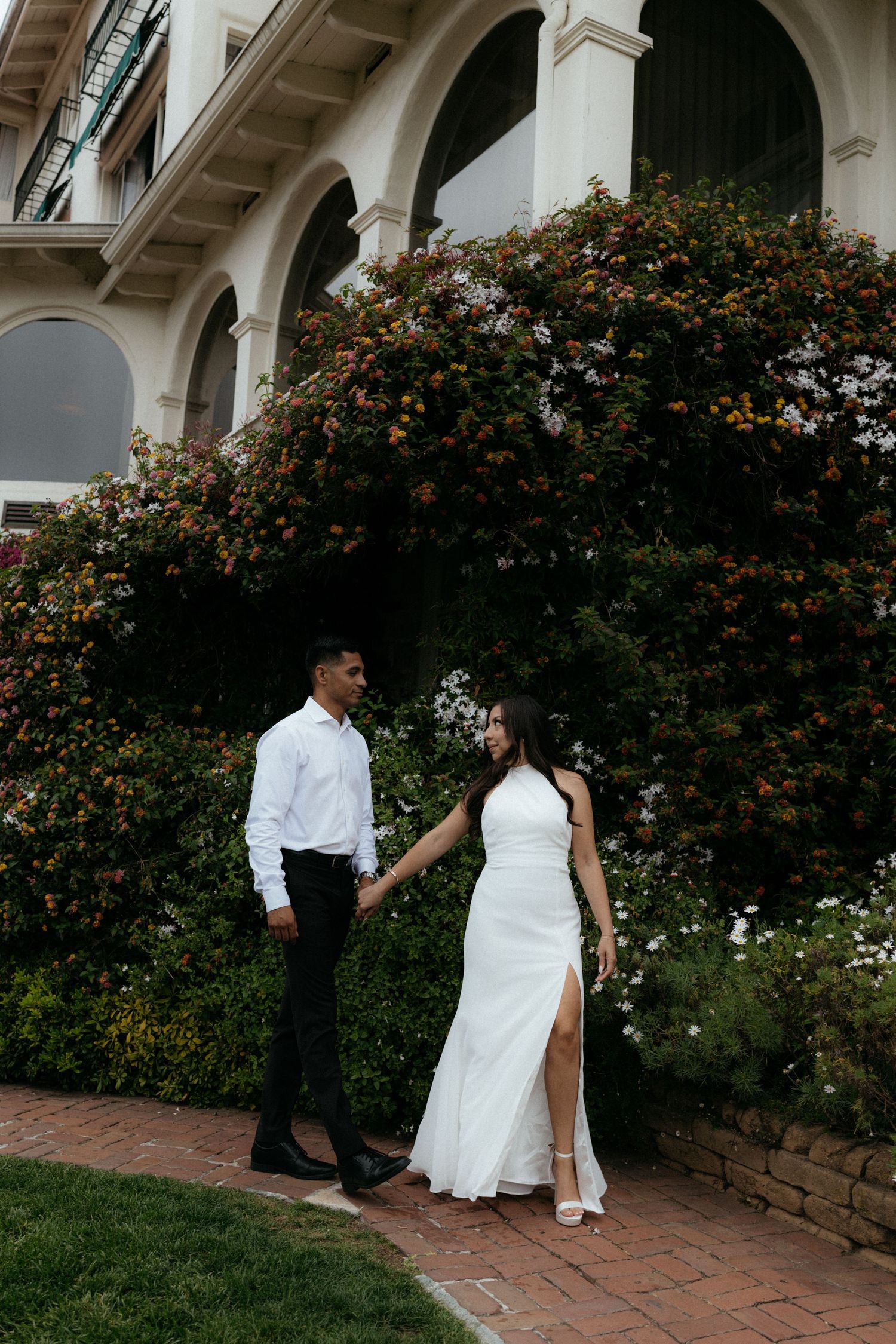 A couple in wedding attire holds hands on a brick path in front of a flowering bush at an elegant venue.
