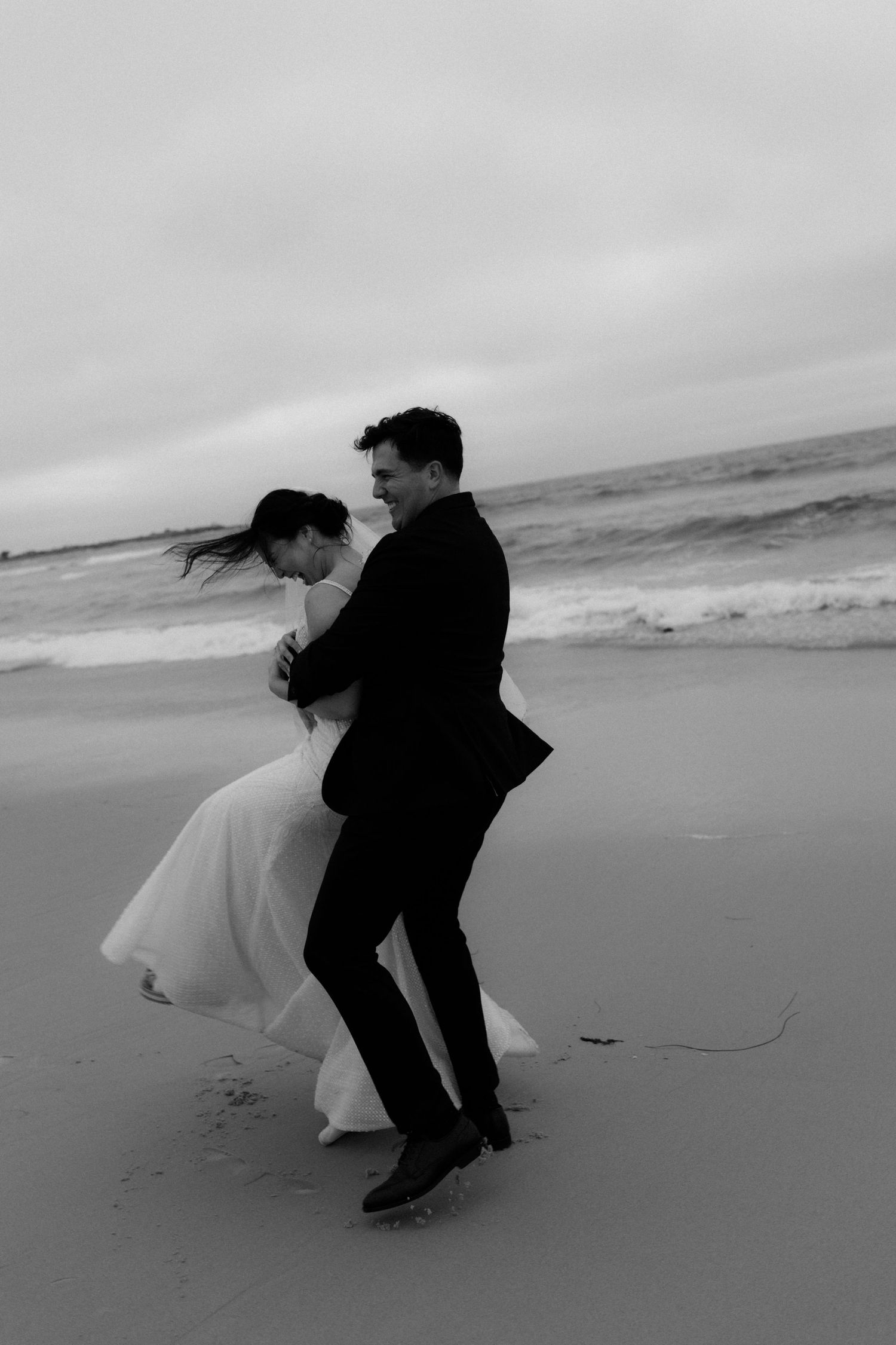 Black and white photo of a joyful couple dancing on a windy beach in wedding attire, waves crashing behind them.