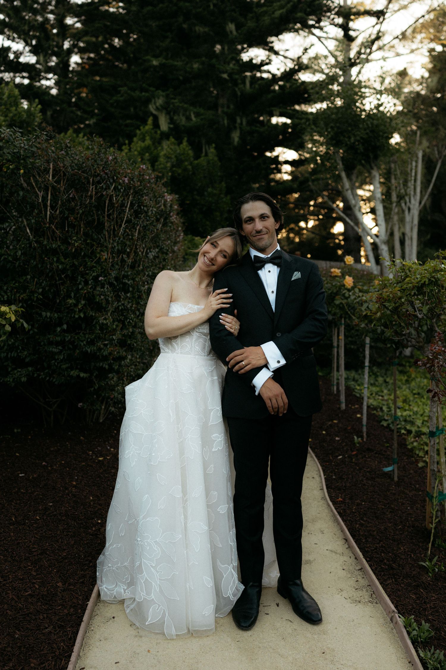 Bride in strapless white gown and groom in black tuxedo posing together on garden path at outdoor wedding.