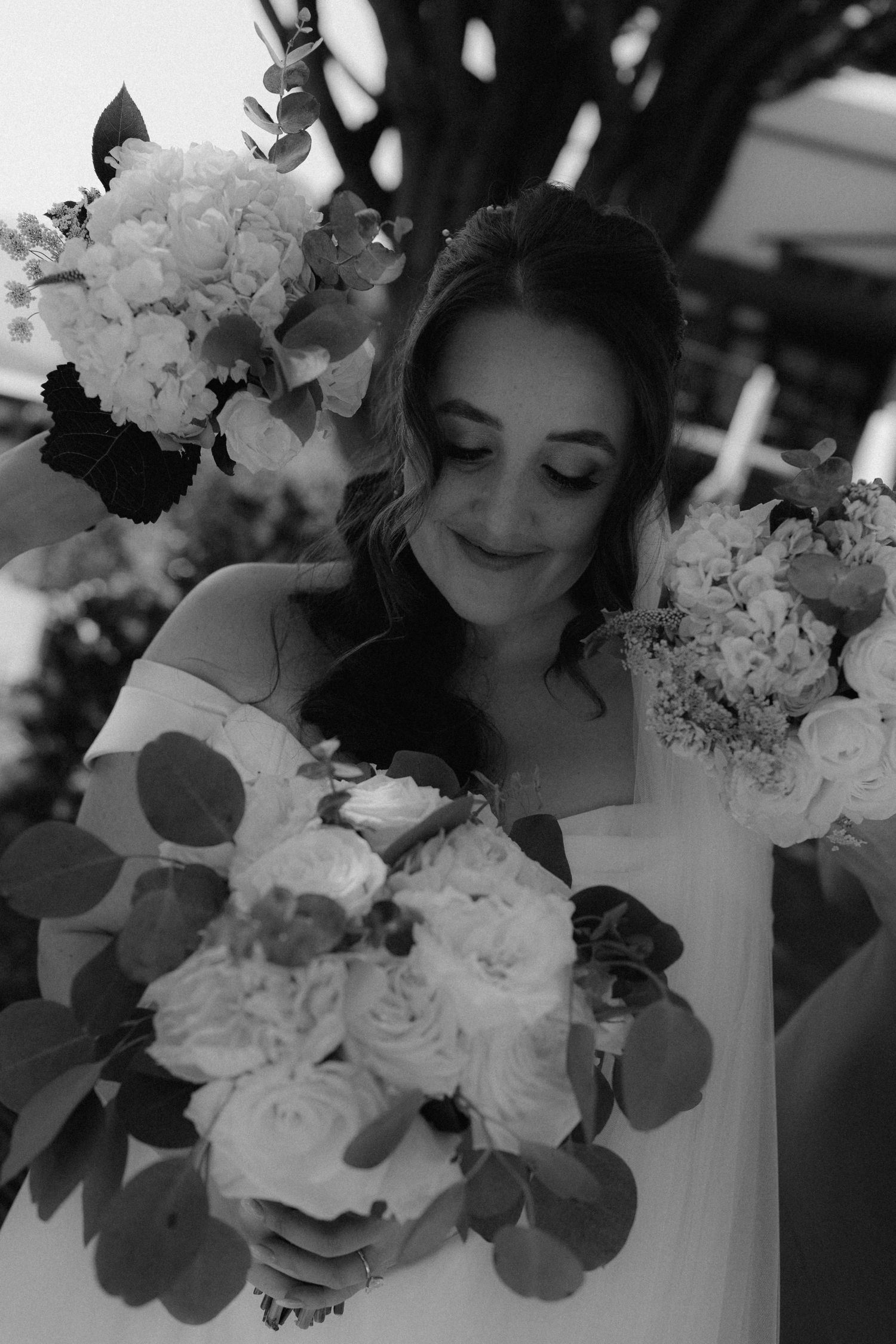 Bride in white gown holds lush bouquets of roses and hydrangeas, smiling softly in black and white photo.
