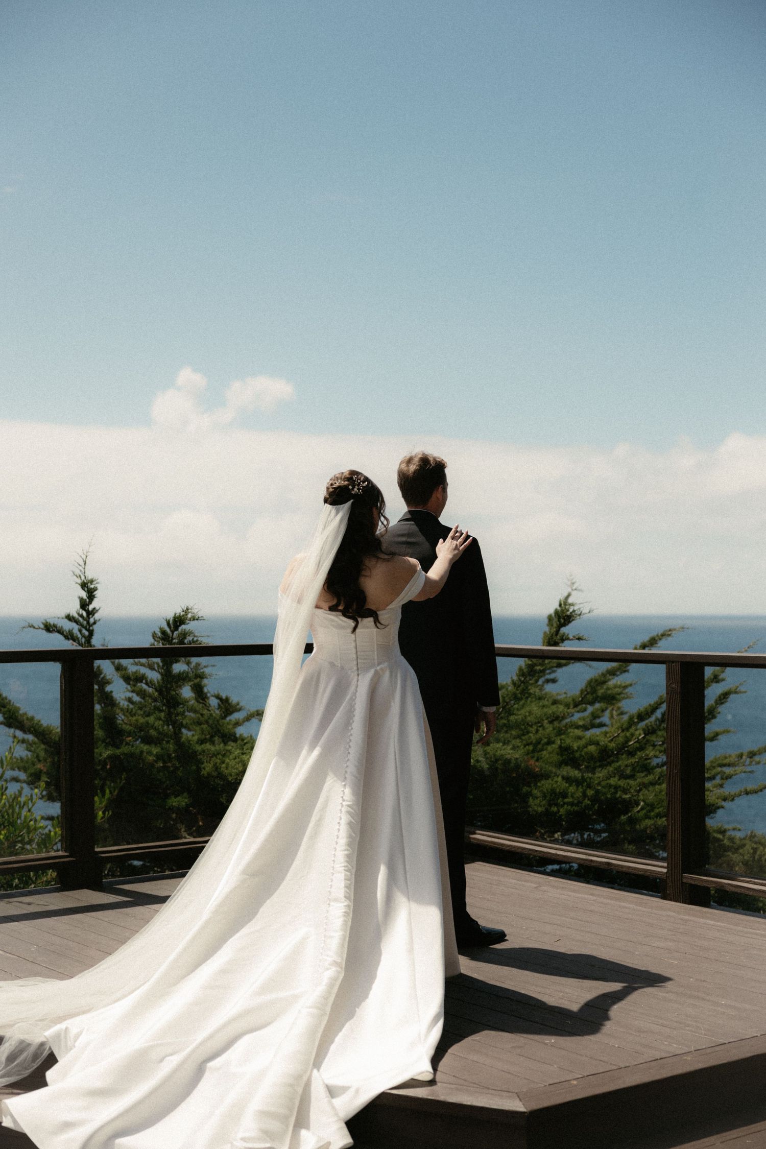 Bride and groom stand on scenic overlook deck, gazing at ocean view, bride in flowing white gown with long veil.