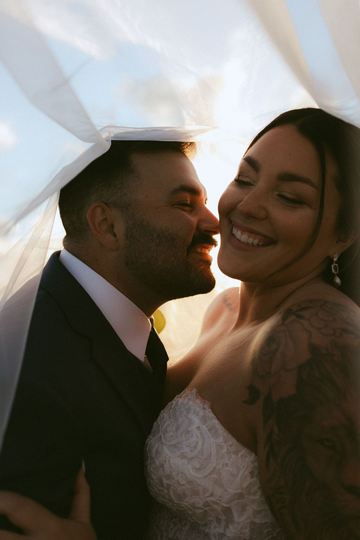 Bride and groom share a romantic moment under a veil, smiling closely together in warm golden sunlight.