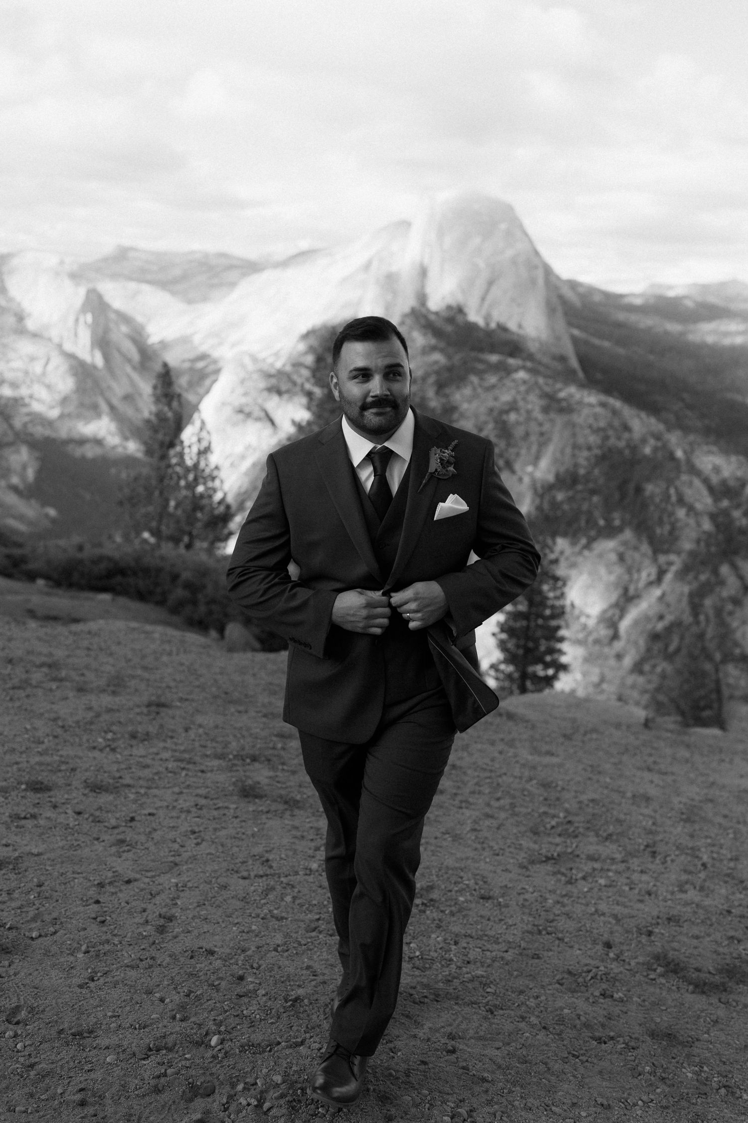Groom in dark suit with boutonniere walks confidently at Yosemite with Half Dome in background, black and white photo.