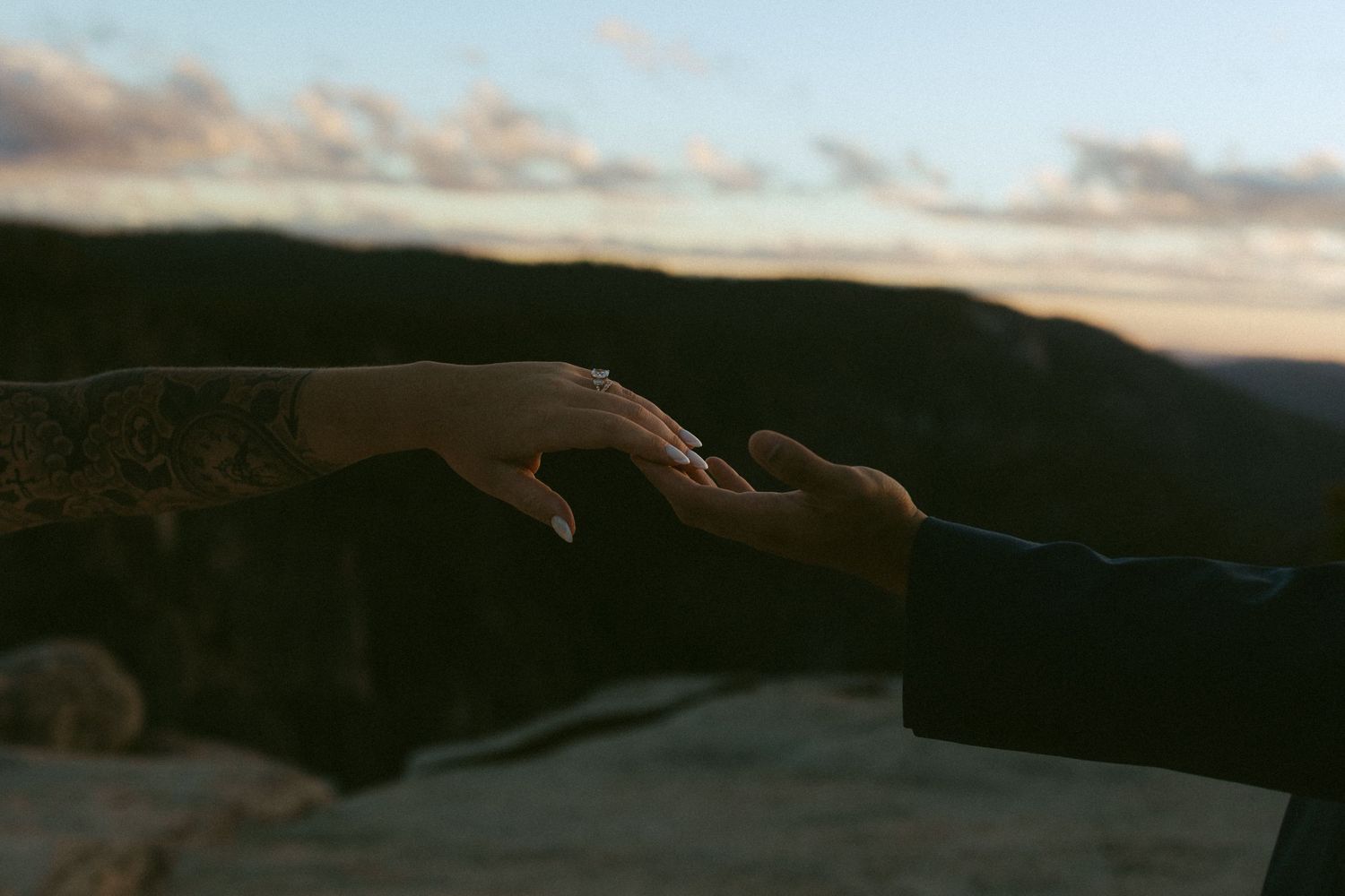 Two hands reaching toward each other at sunset with mountain silhouettes in the background.