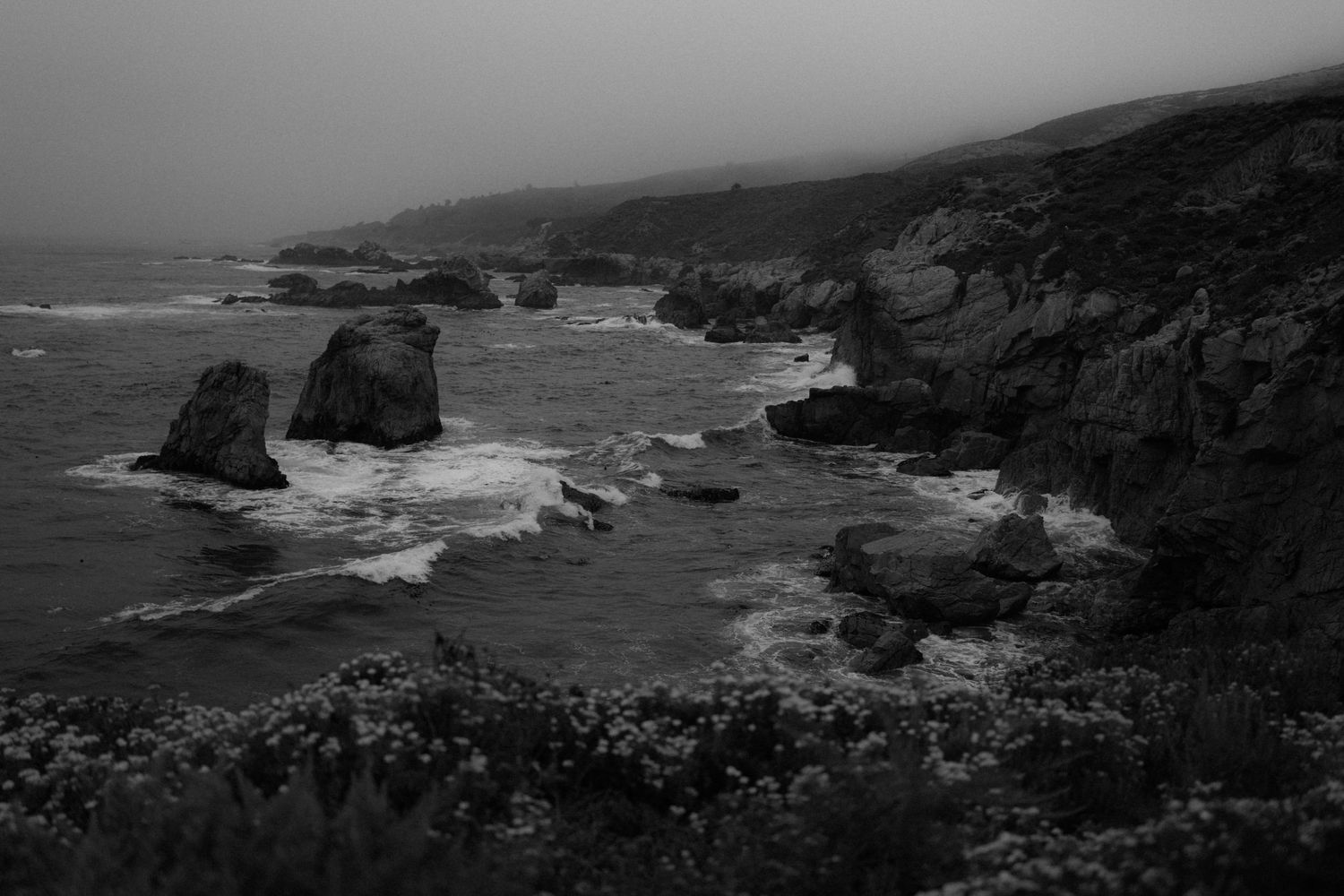 Dramatic black and white coastal landscape with rugged cliffs and crashing waves along a rocky shoreline.