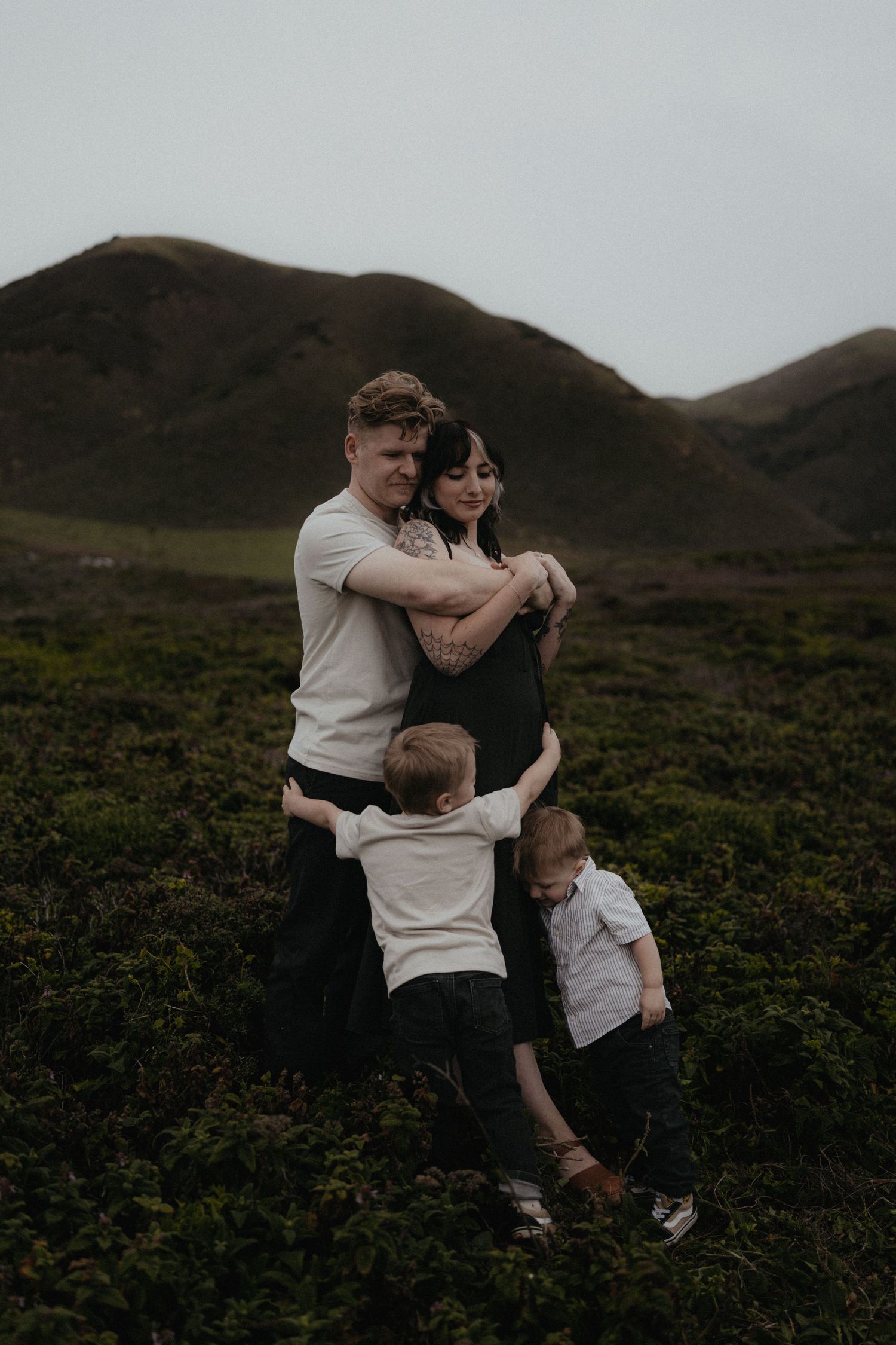 A loving family of four embracing in a green field with scenic mountain backdrop on overcast day.
