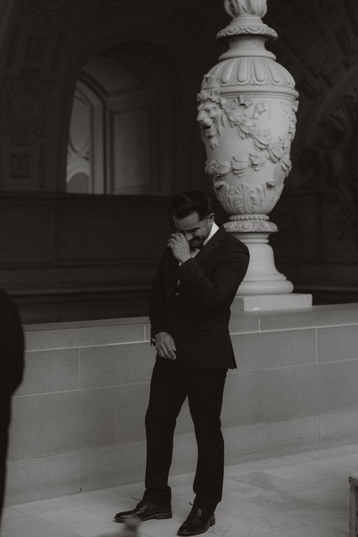 Black and white photo of a groom in a suit standing near ornate urn column, looking down emotionally.