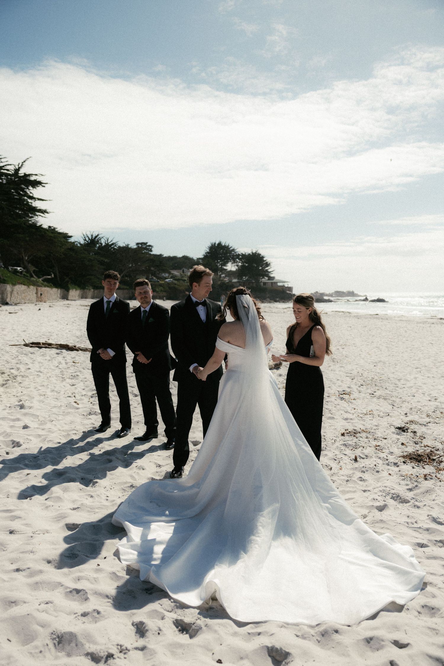 Bride and groom exchange vows on sandy beach with groomsmen and officiant under blue sky.