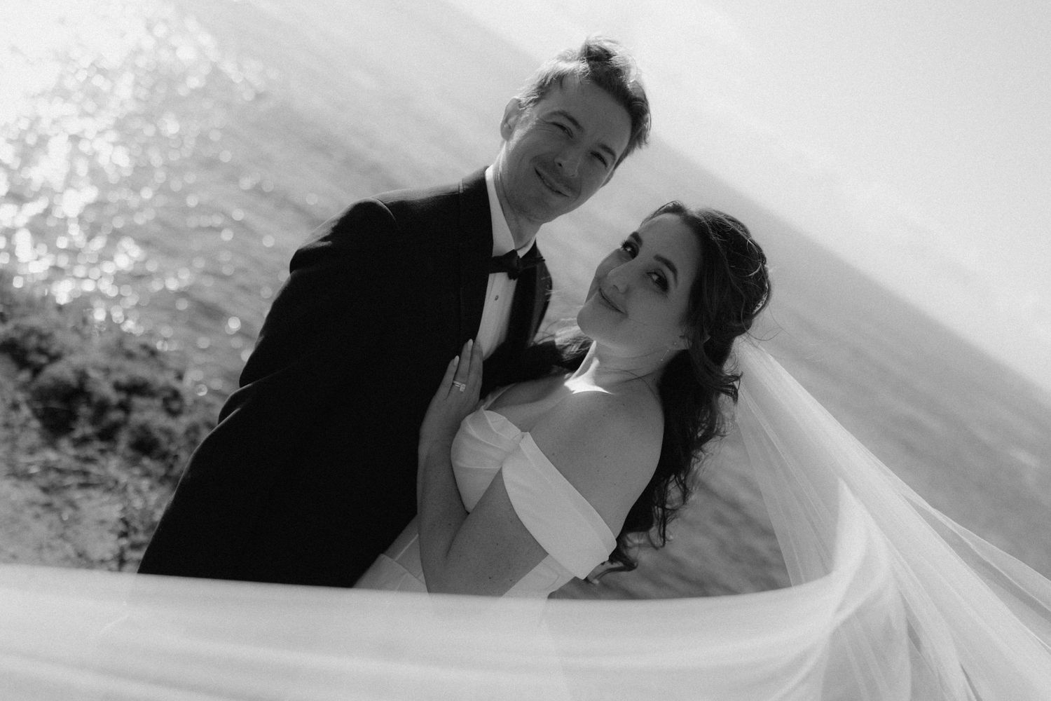 Black and white wedding photo of bride and groom posing by the ocean, veil flowing in the wind.