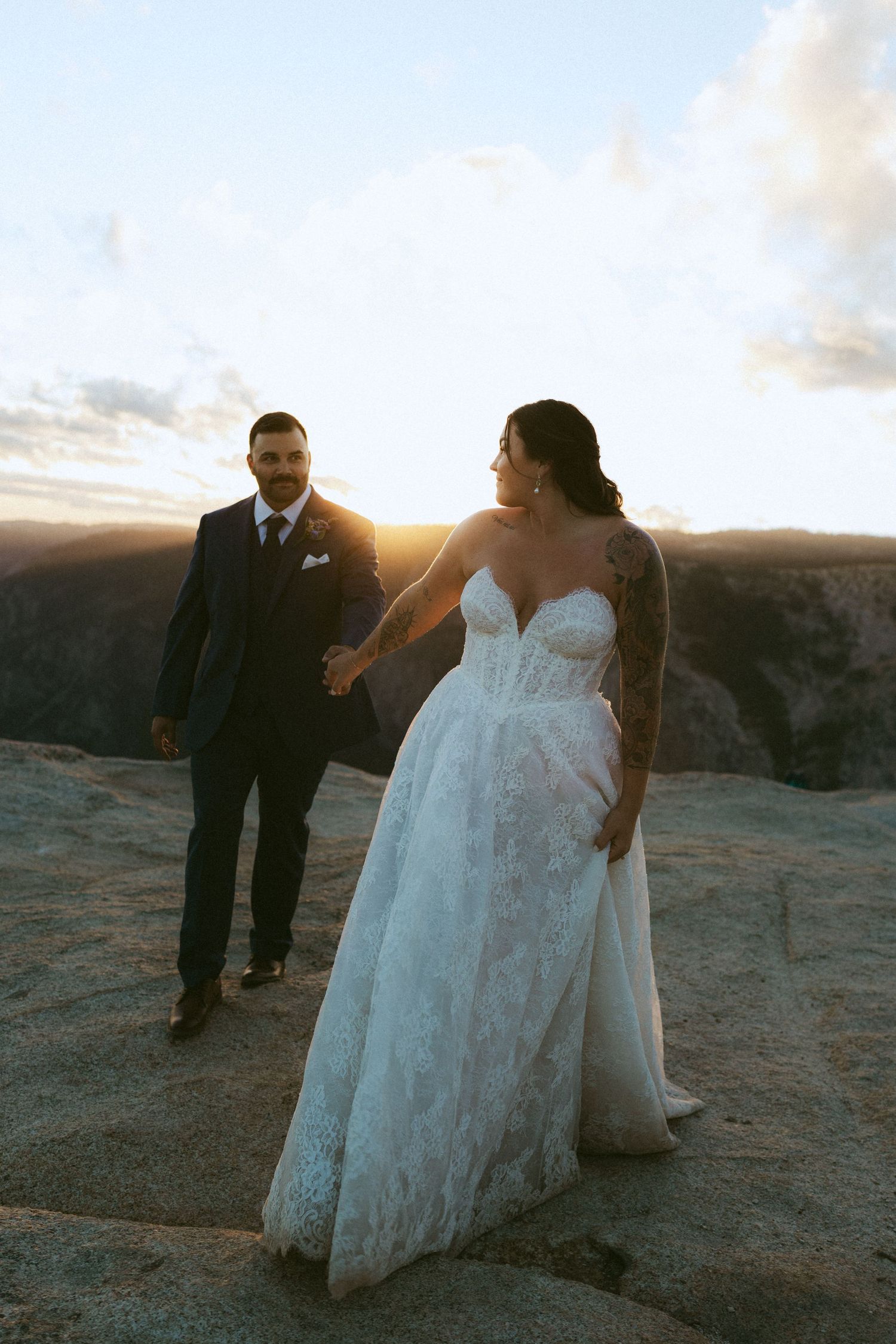 Bride and groom hold hands at sunset on a rocky cliff, bride wearing lace gown with floral tattoo sleeve.