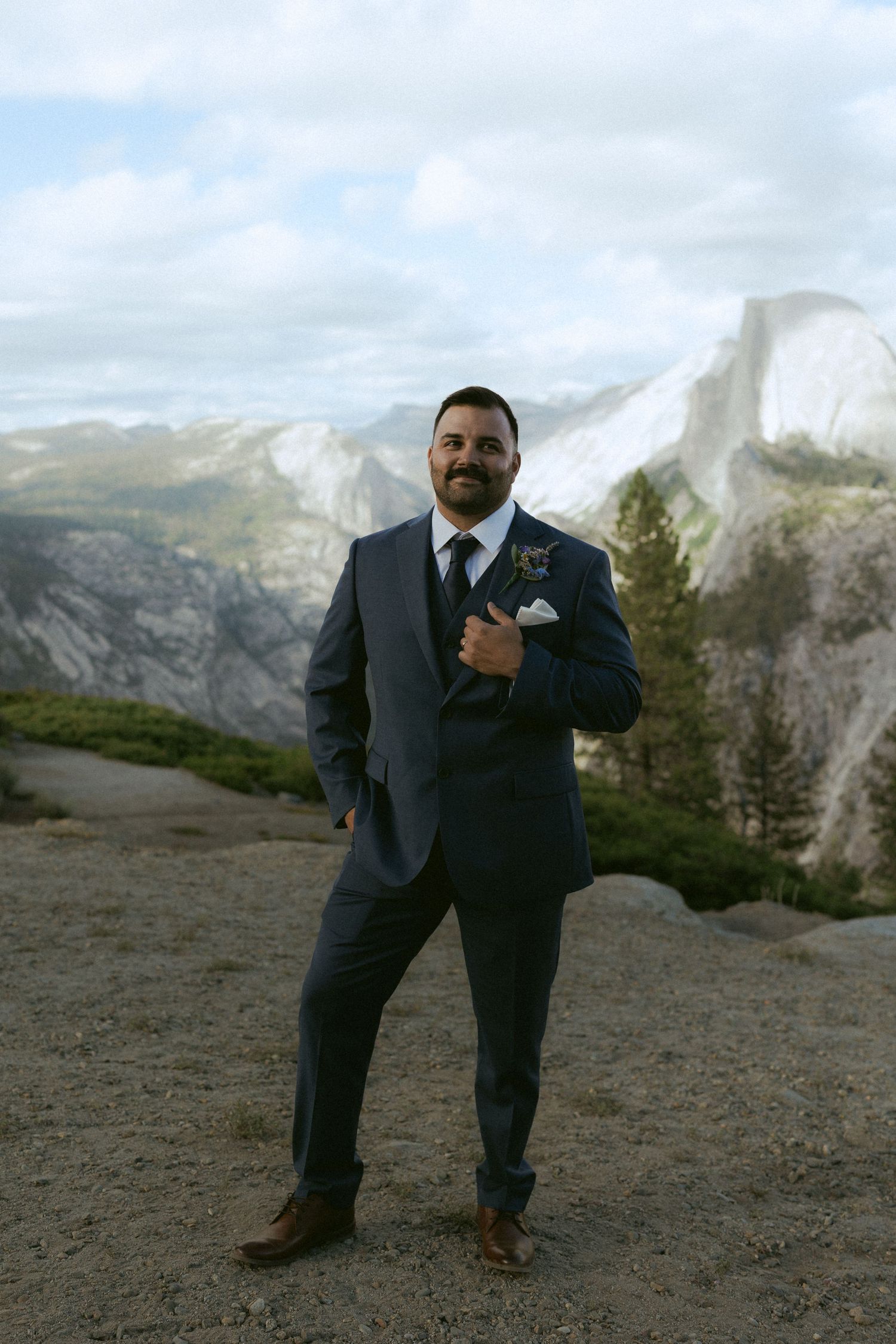 Smiling groom in navy suit poses confidently at Glacier Point with Half Dome visible behind him.