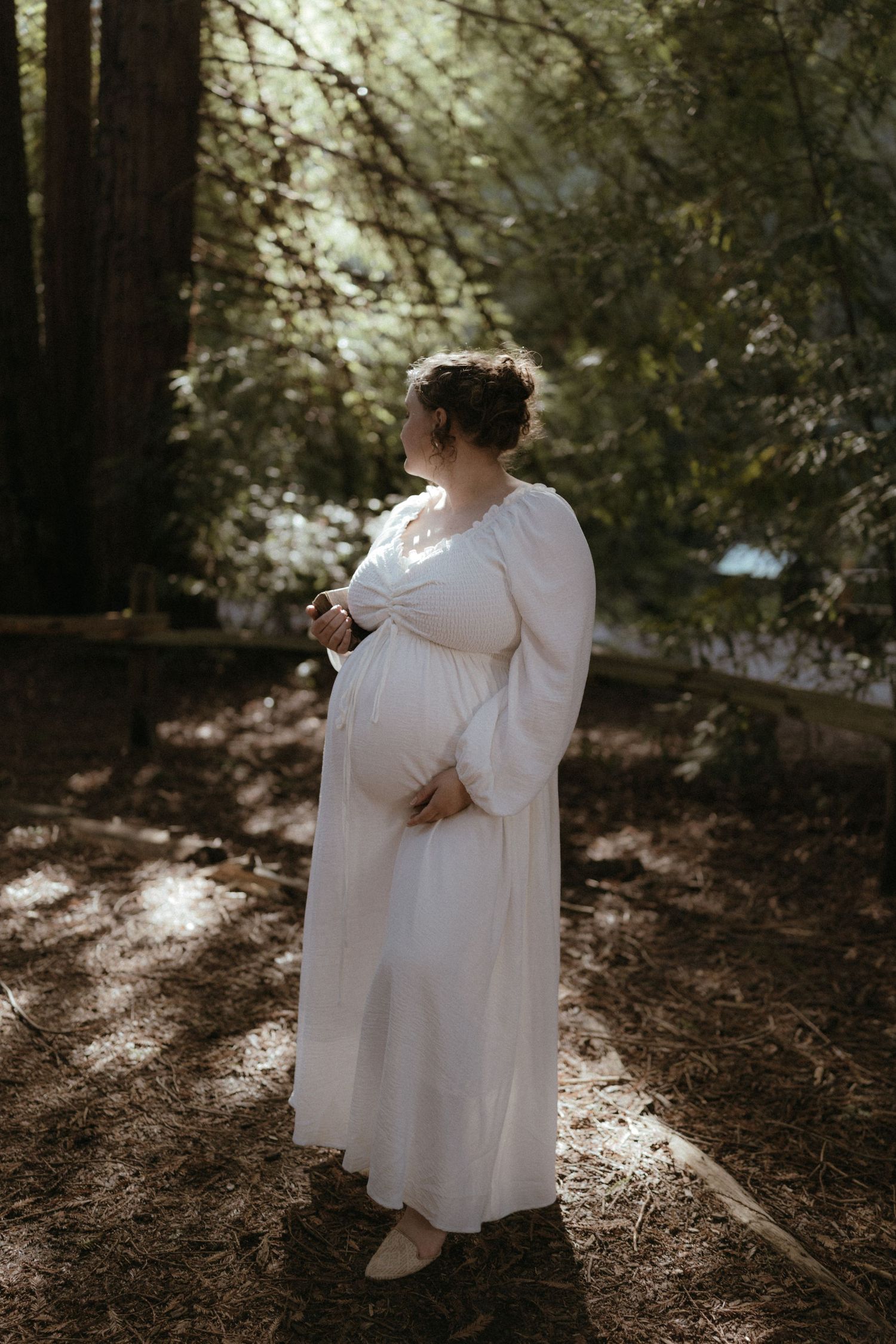 A person in a flowing white dress stands in dappled forest sunlight with hands on their belly.
