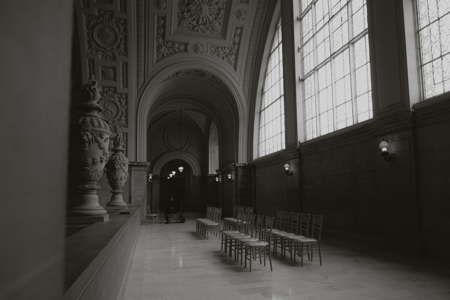 An elegant hallway with ornate architecture, tall windows, and rows of chairs.
