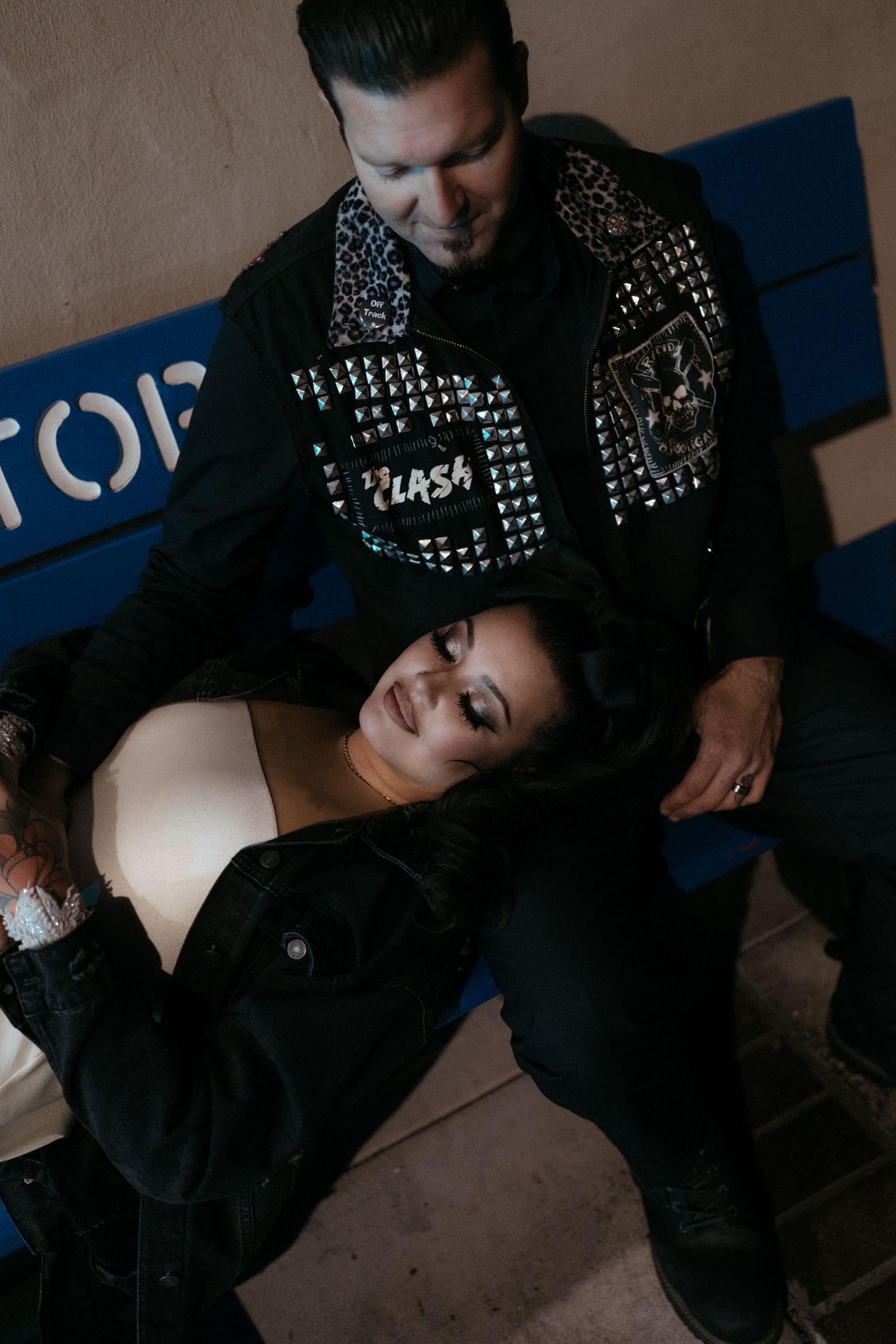 A couple in contrasting formal and punk attire pose together in dramatic lighting.