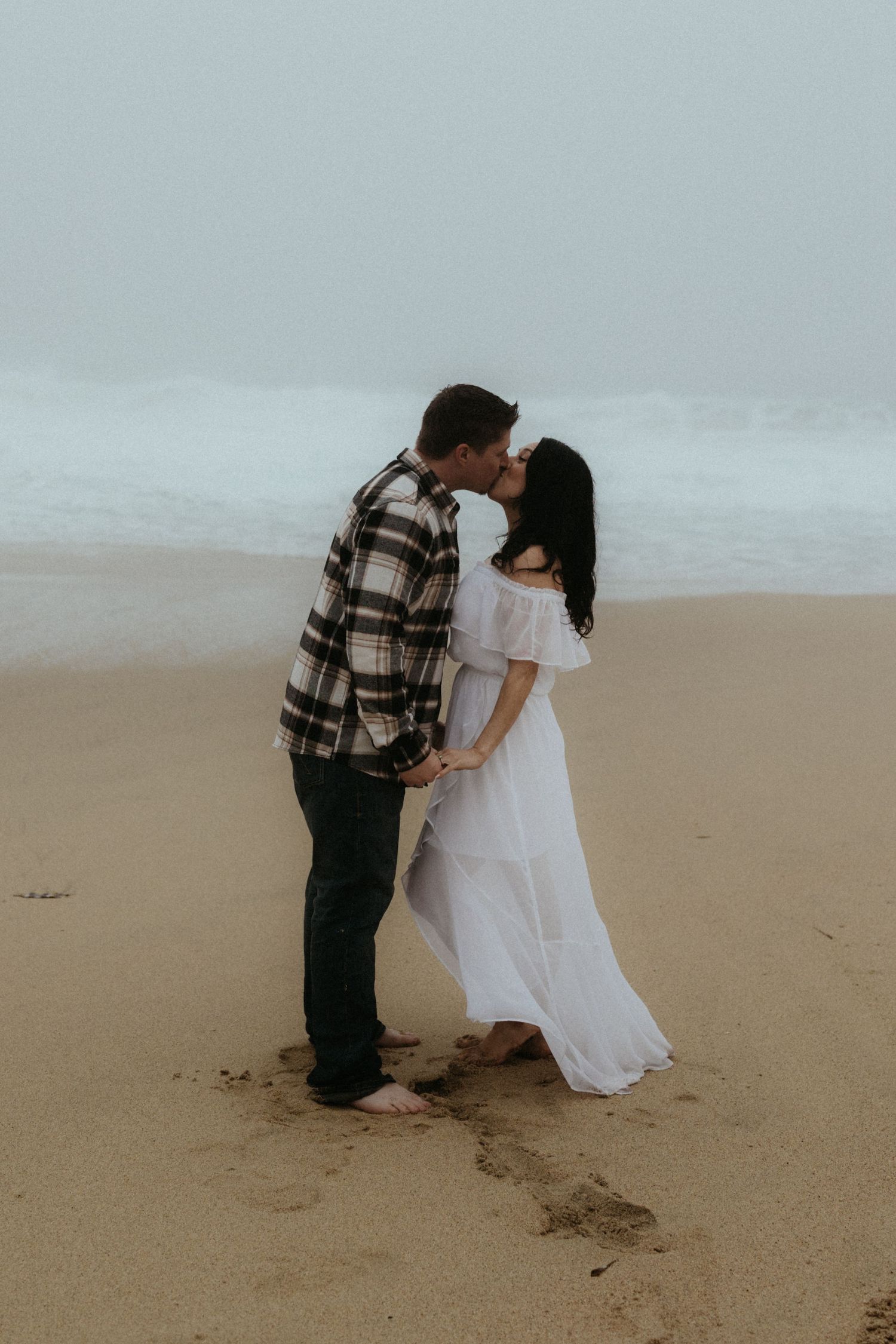 Couple in white dress and plaid sharing moment on misty beach.