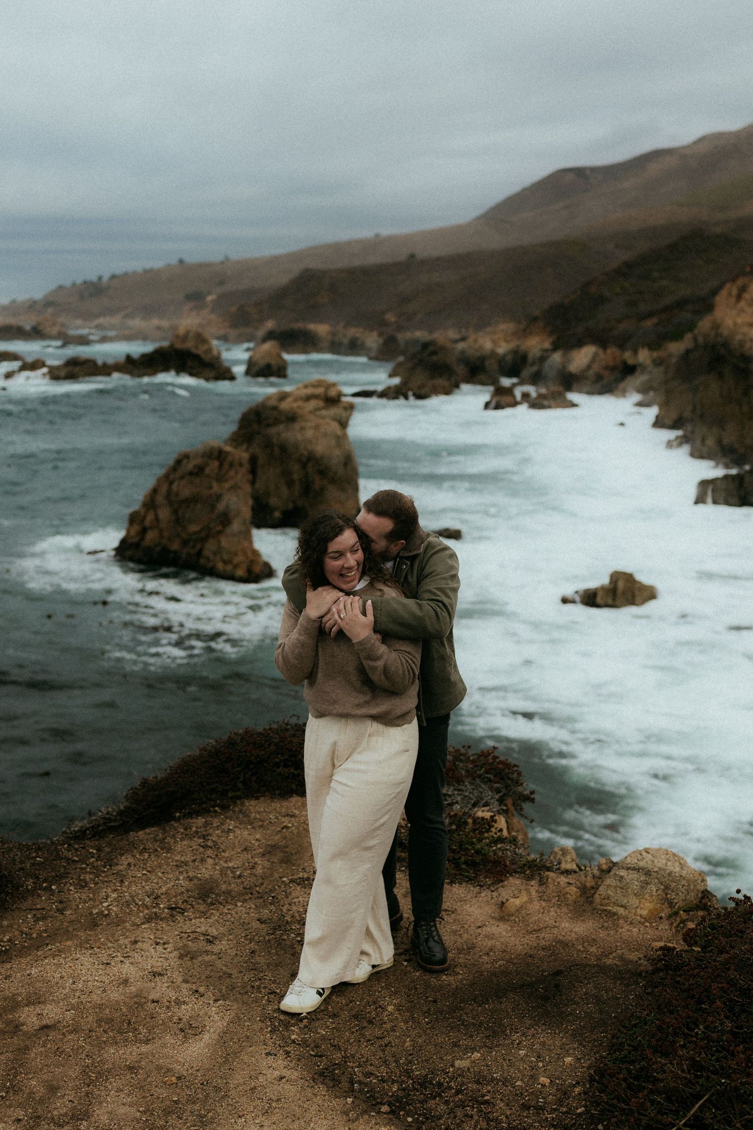 Couple embraces on dramatic coastal cliffs overlooking crashing waves and rocky shoreline at sunset.