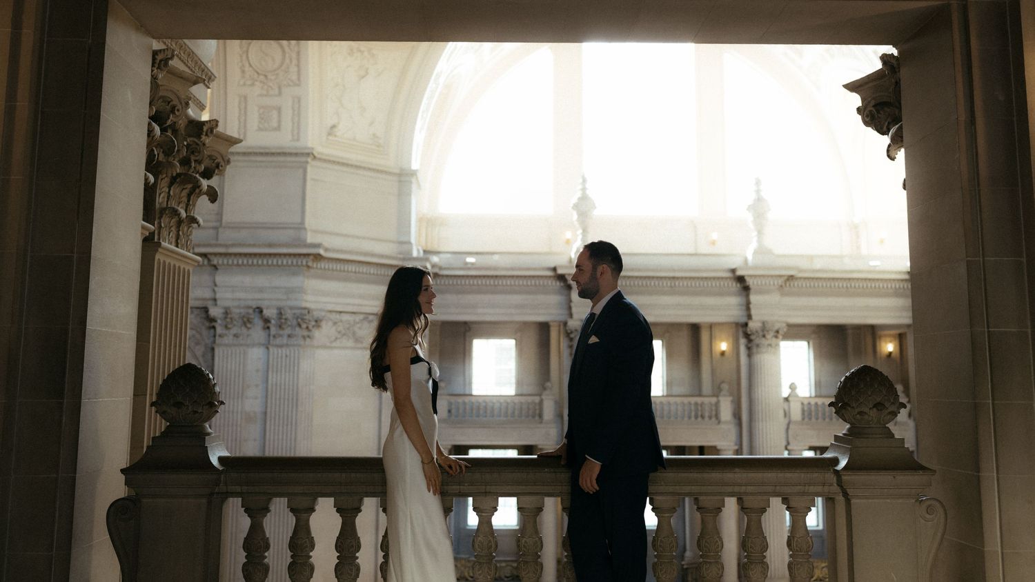 A couple embraces in a grand ballroom with ornate architectural details and dramatic lighting.