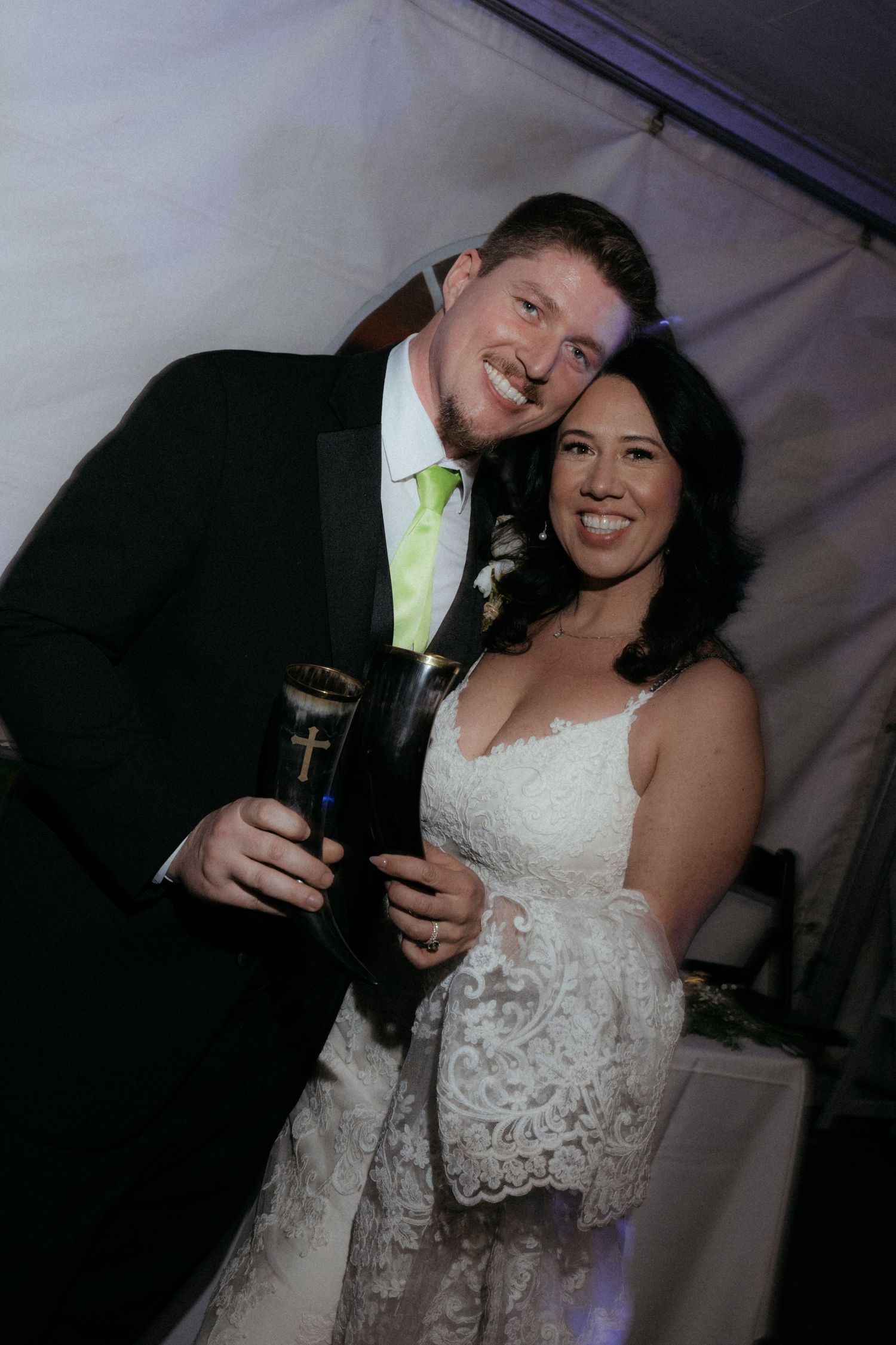A newlywed couple celebrates with drinks at their wedding reception under a tent.
