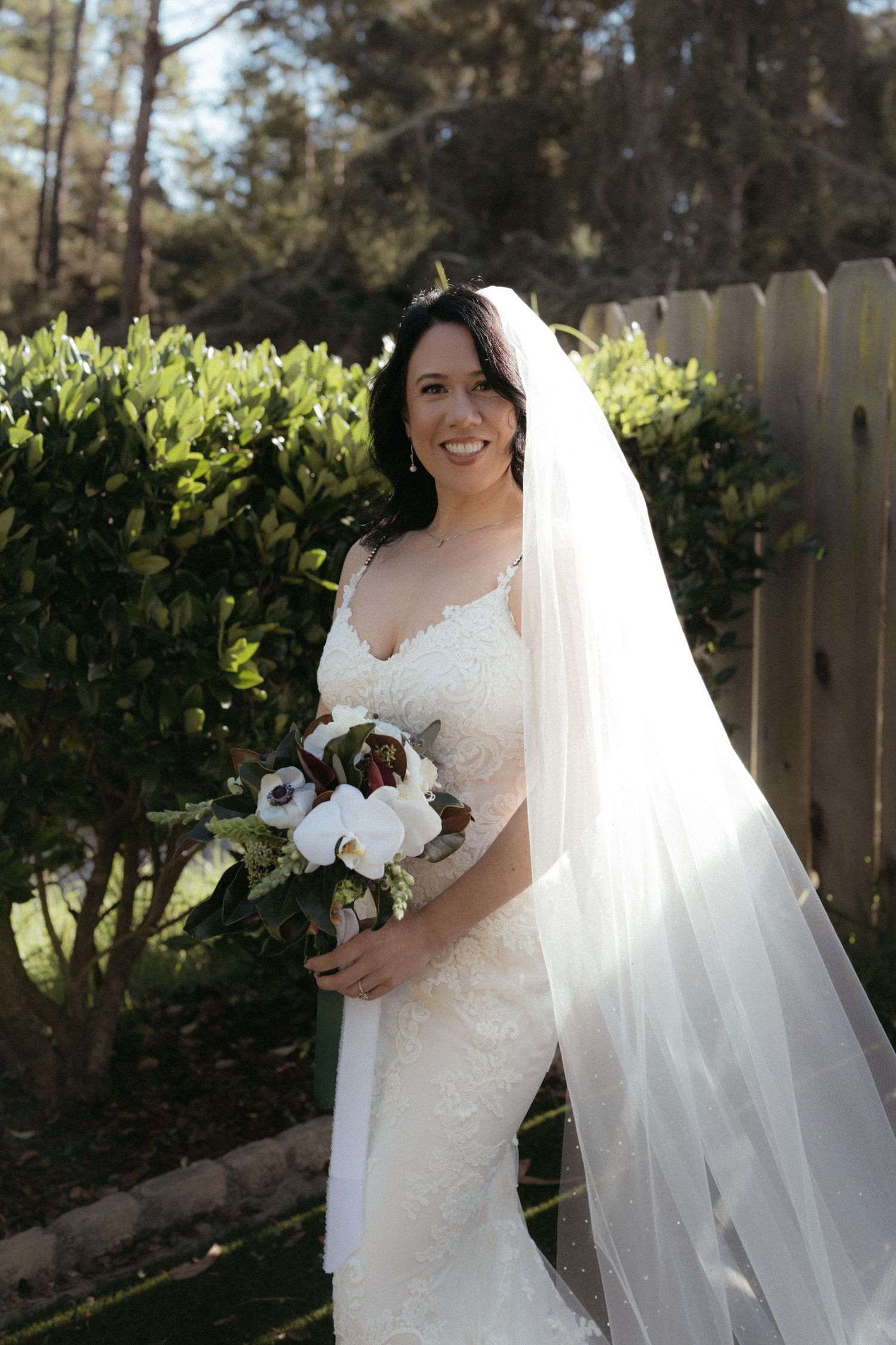 A bride in a white lace wedding dress and veil poses outdoors with a white floral bouquet in a garden setting.