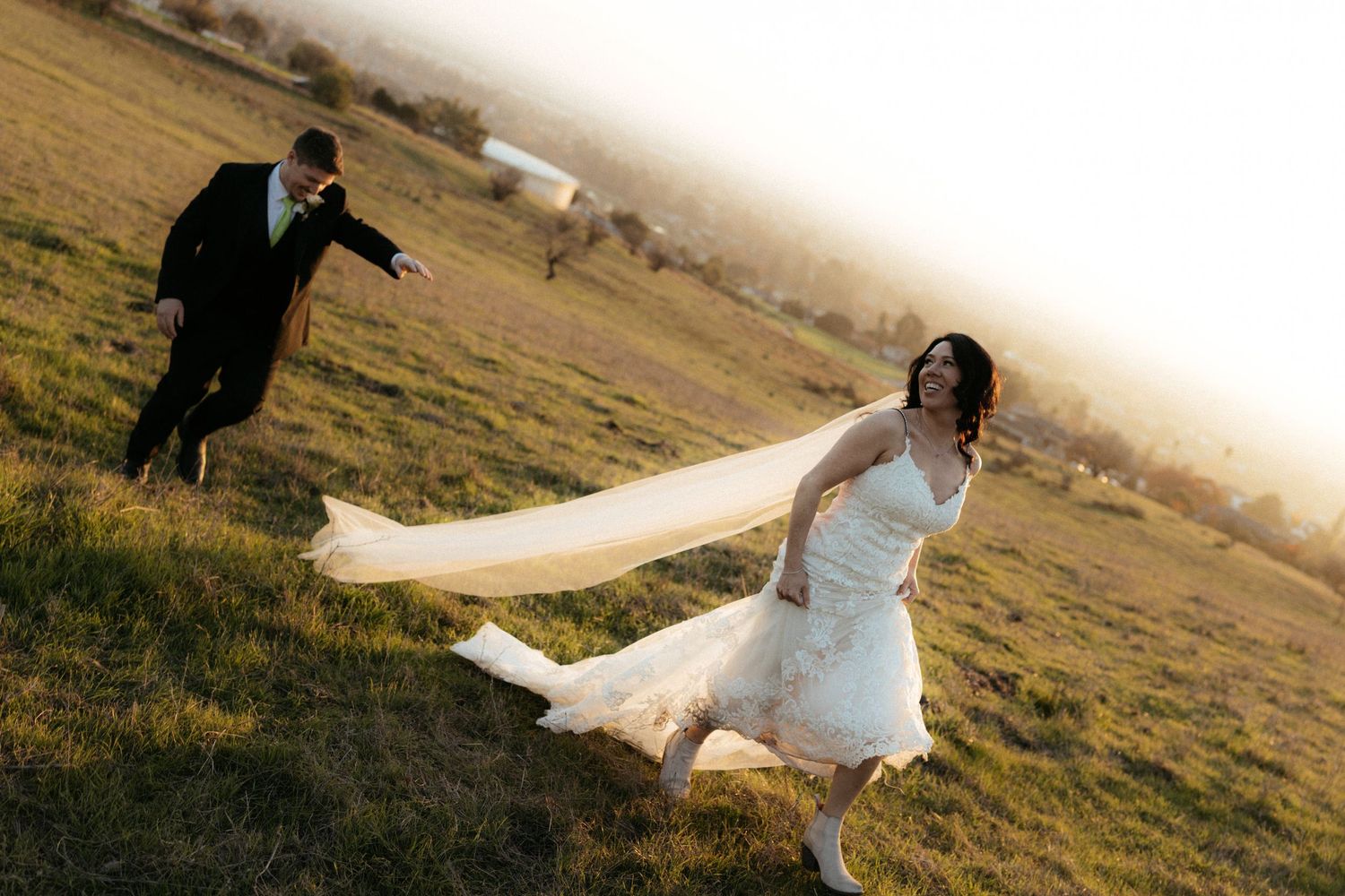 A playful wedding couple runs through a grassy field with the bride's veil flowing in the wind.