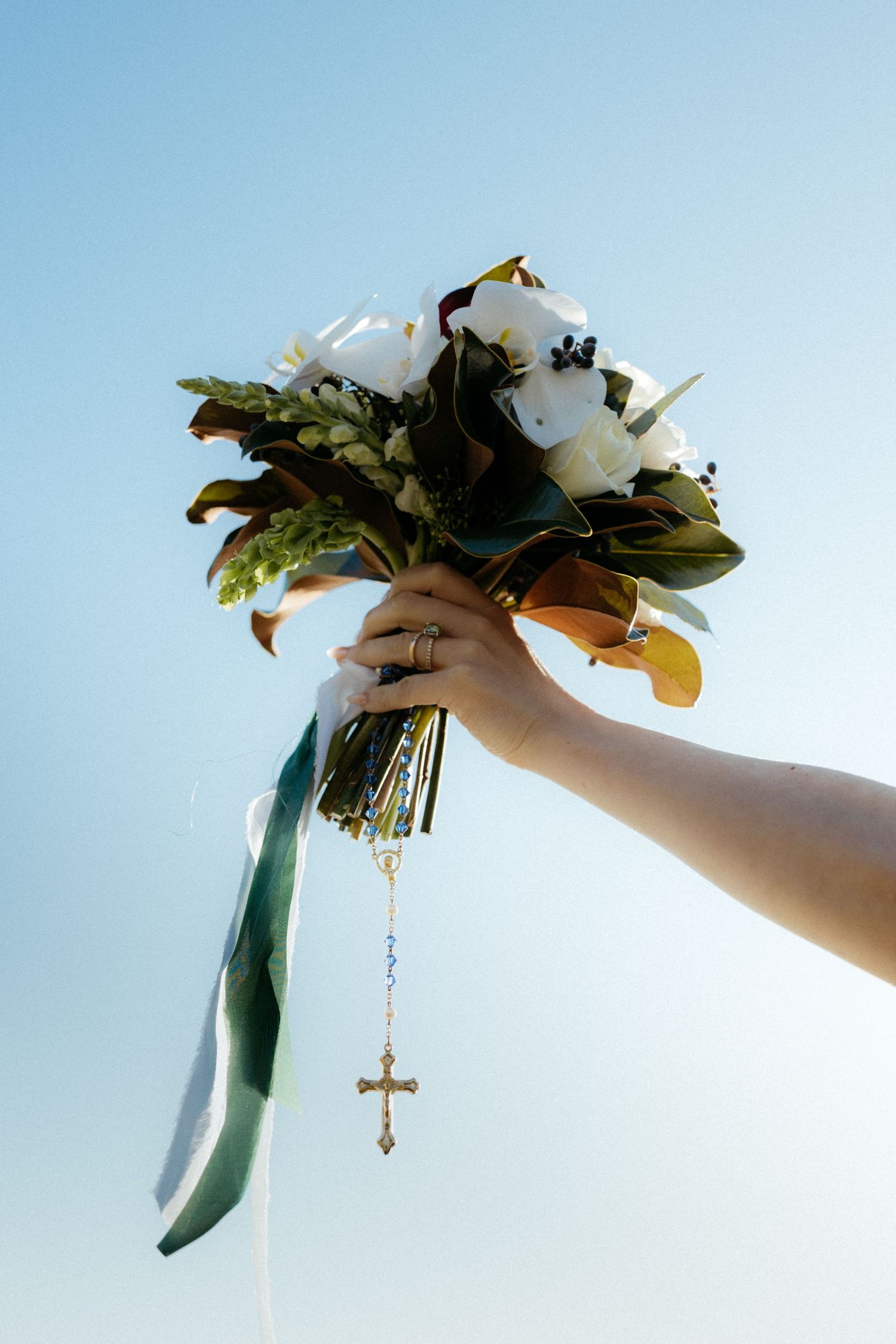 A hand holds up a delicate white floral bouquet with greenery against a soft blue sky background.