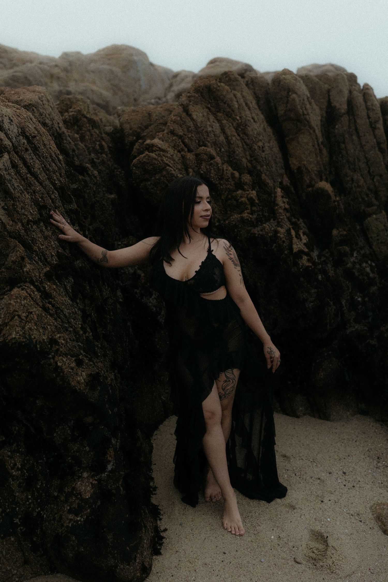 Person in dark clothing poses artistically against rocky beach backdrop.