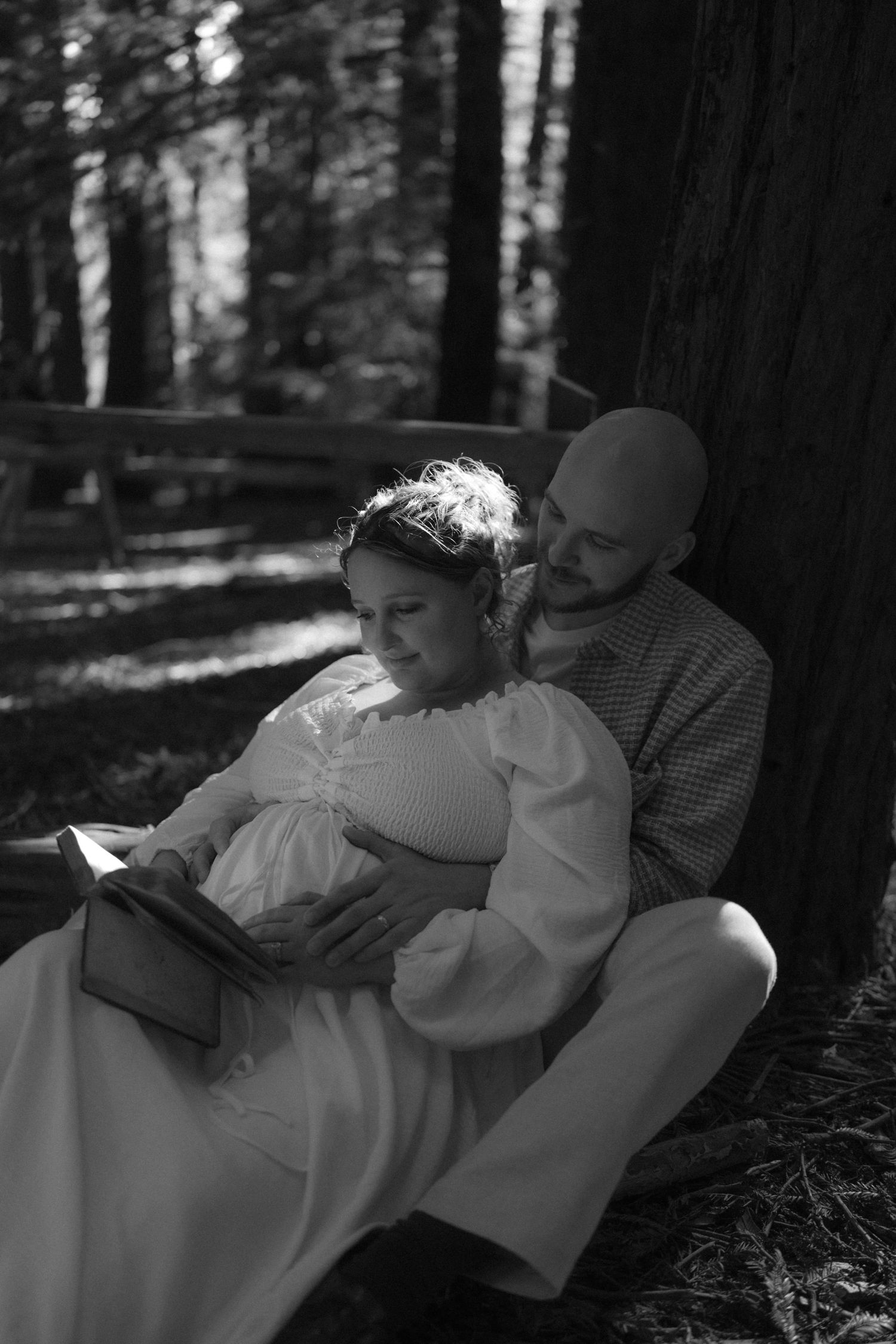 A couple shares an intimate moment while resting against a tree in a black and white forest scene.