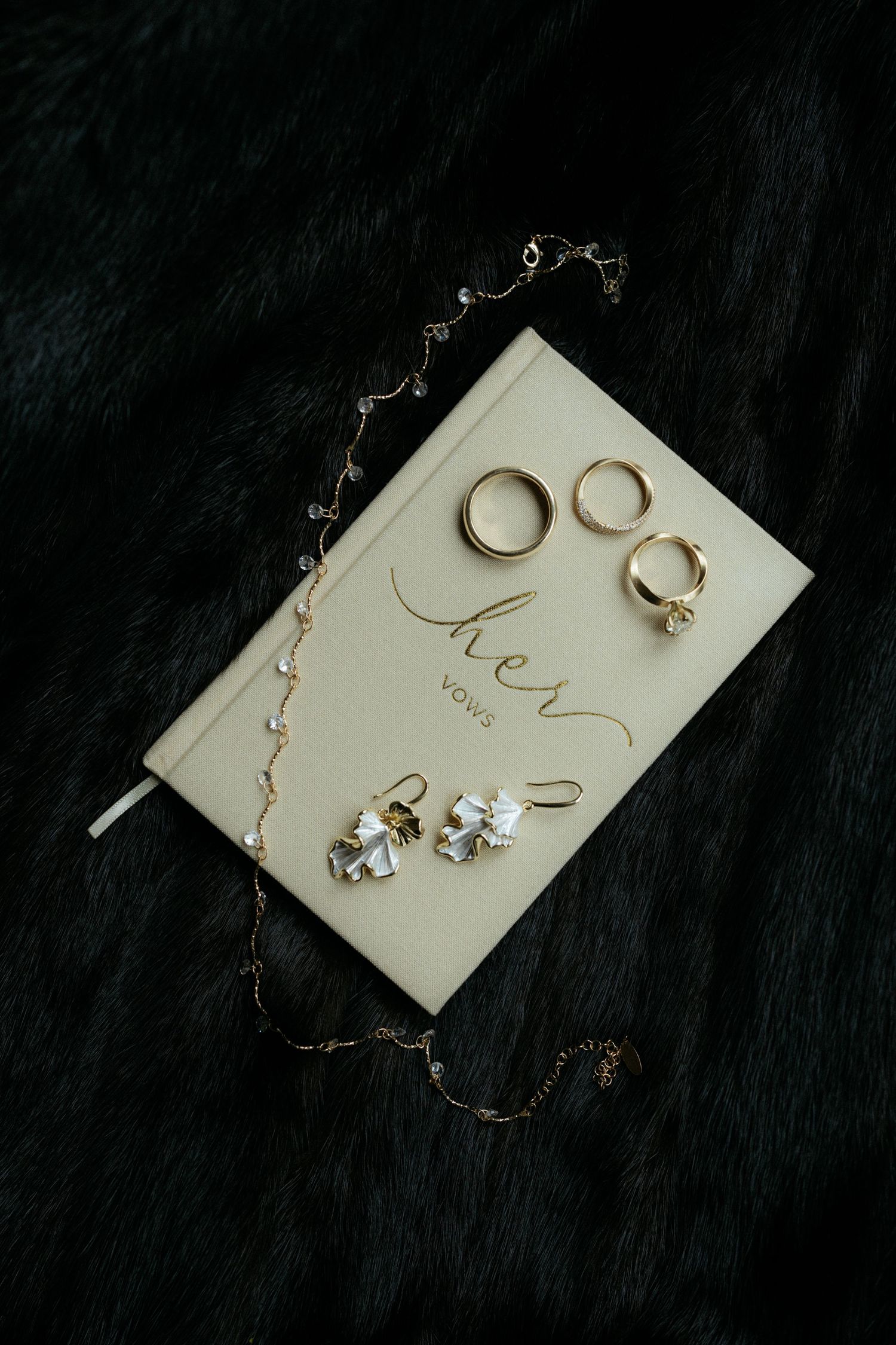 Gold wedding rings, crystal earrings, and a delicate necklace arranged on a white her vows book atop dark fur.