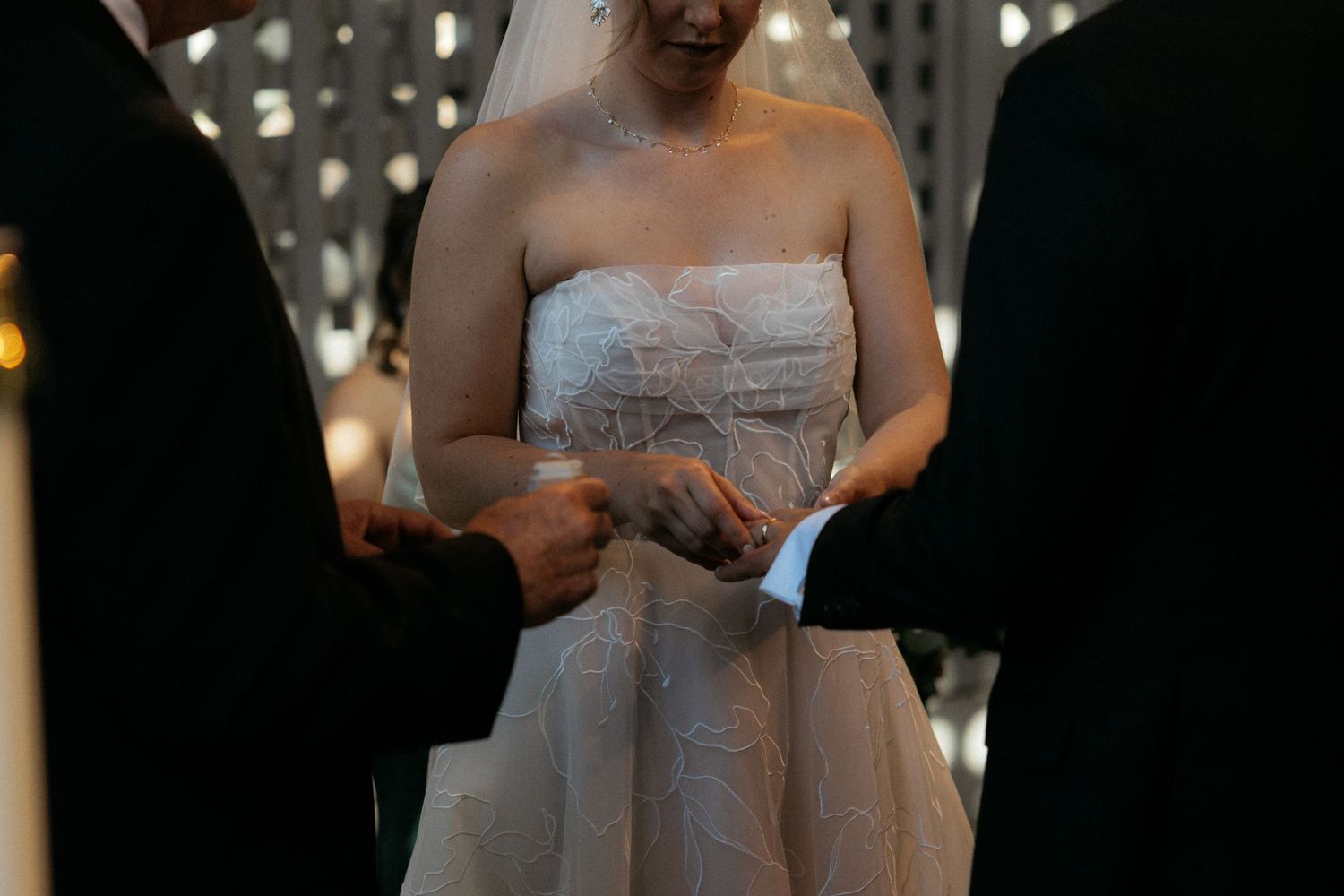 Bride in strapless lace wedding gown exchanging rings with groom during elegant indoor wedding ceremony.