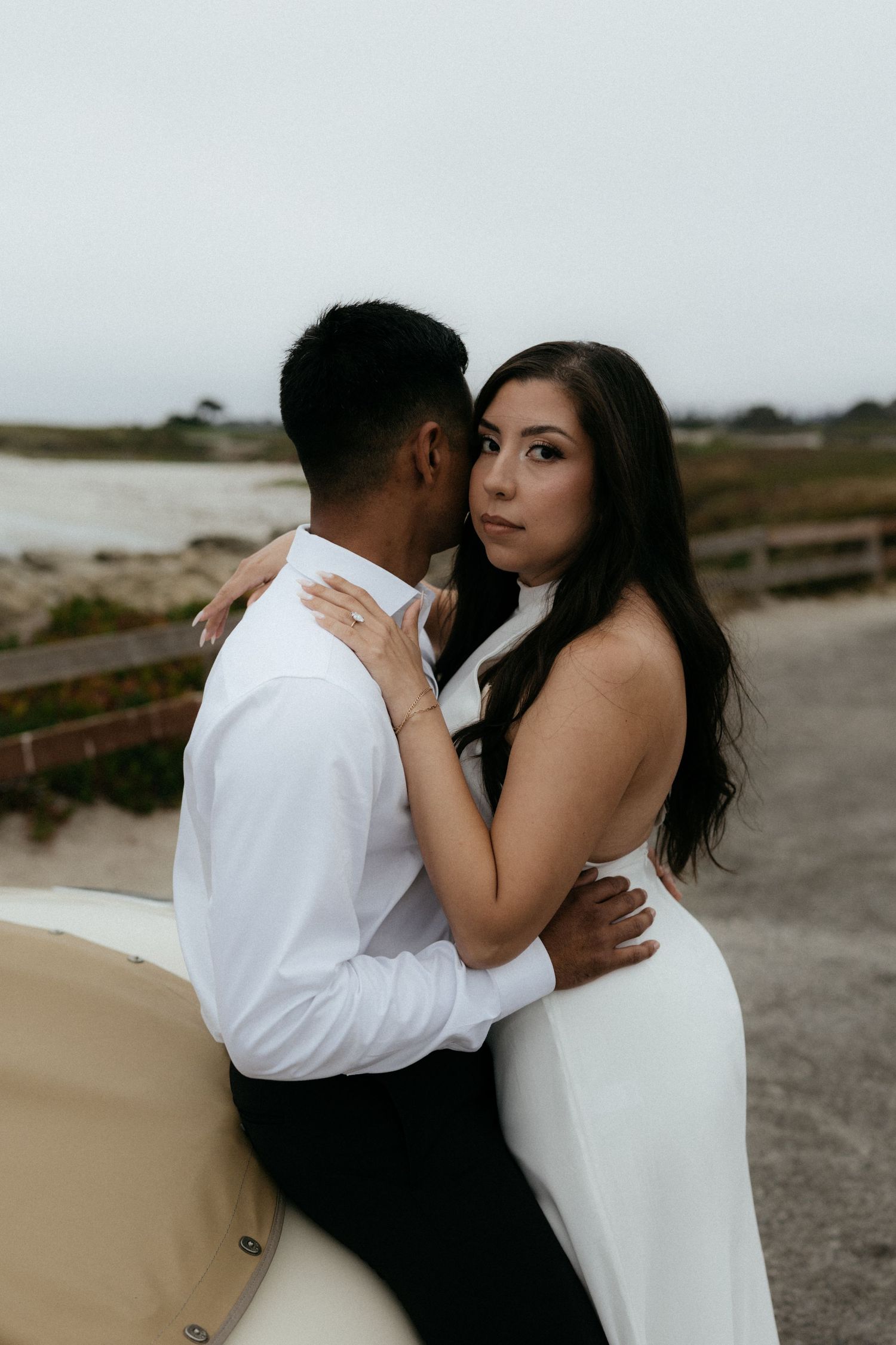 Couple embracing near a vintage car on a misty beach in white attire.