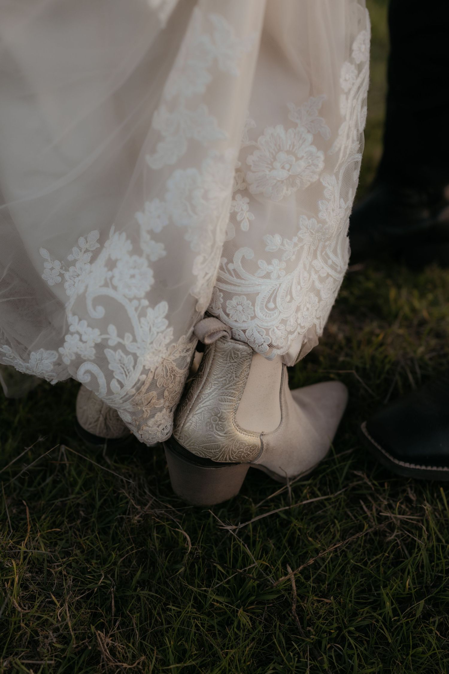 Close-up detail of lace wedding dress hem and boots on grass.