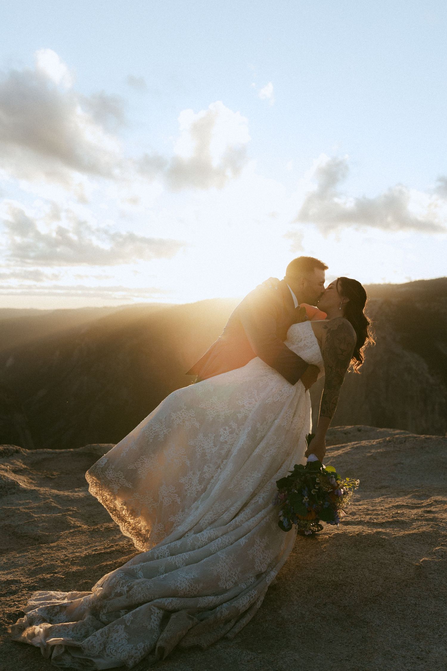 Bride and groom sharing a romantic kiss at sunset on a mountain cliff, wedding dress flowing in the wind.