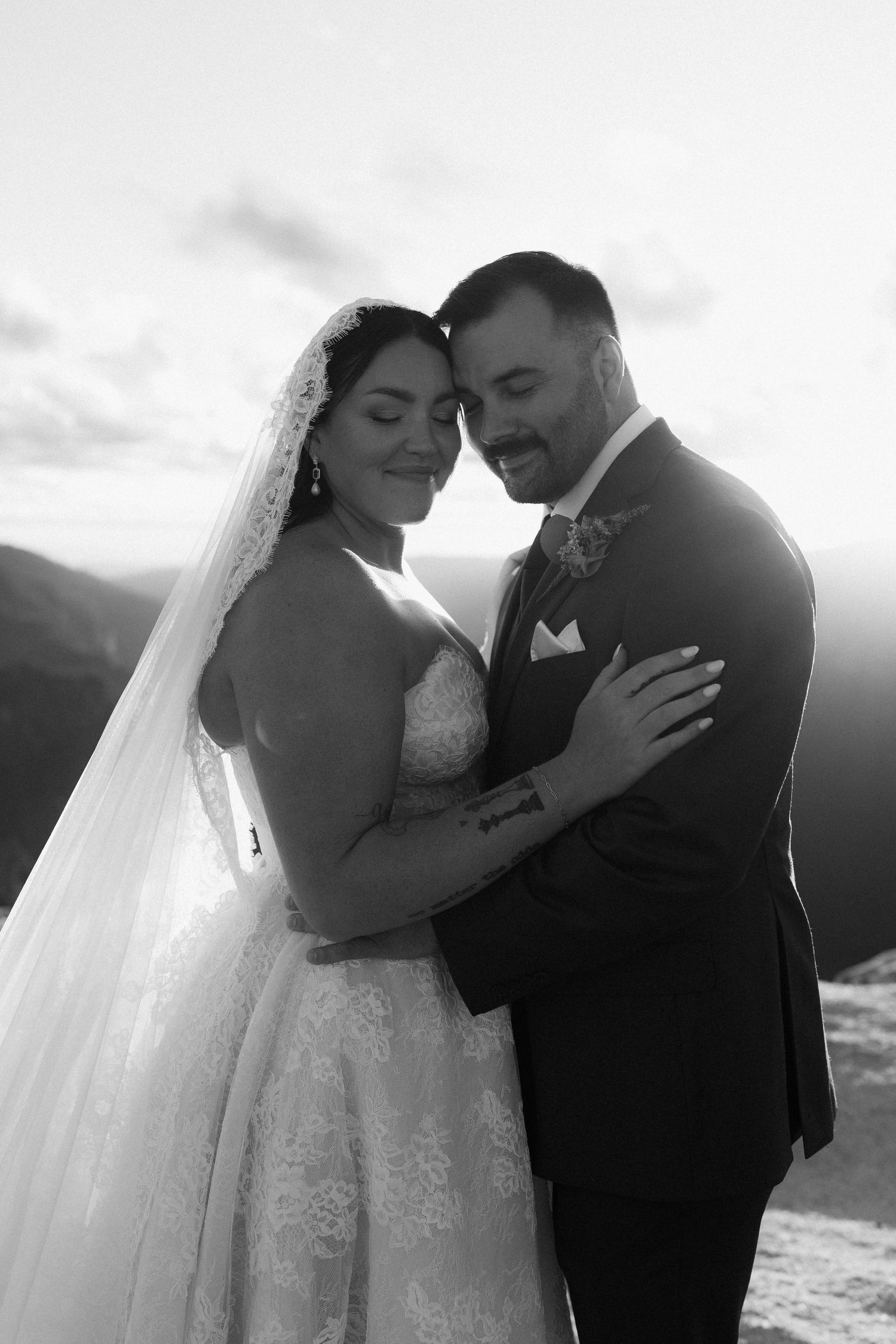 Bride and groom embrace tenderly on a mountain summit, bride in lace gown with veil, black and white portrait.