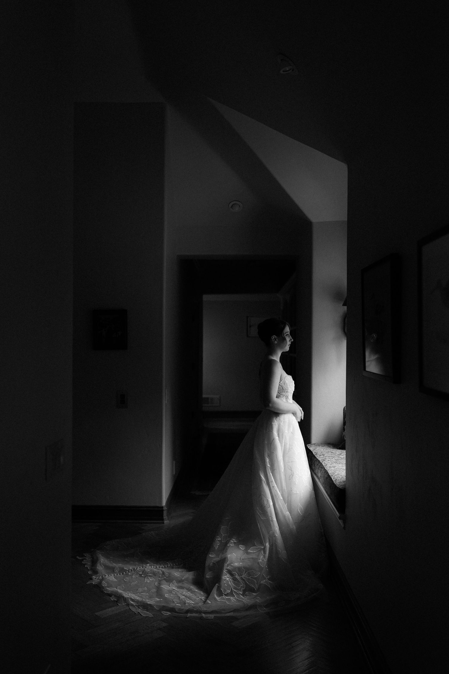 Elegant bride in flowing white gown stands in dramatic shadowed doorway before wedding ceremony.