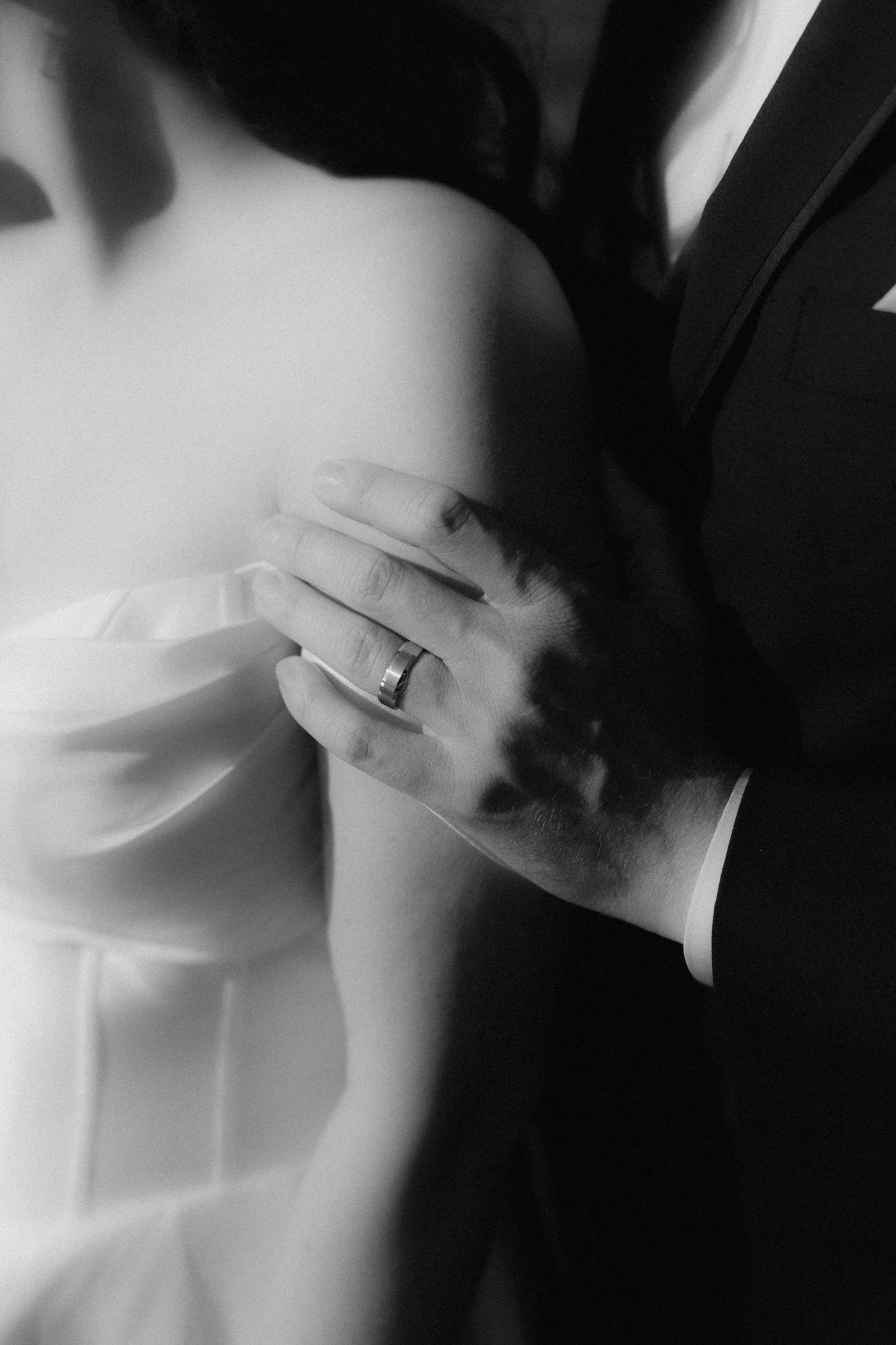 Black and white photo of groom's hand with wedding ring resting on bride's white off-shoulder wedding dress.