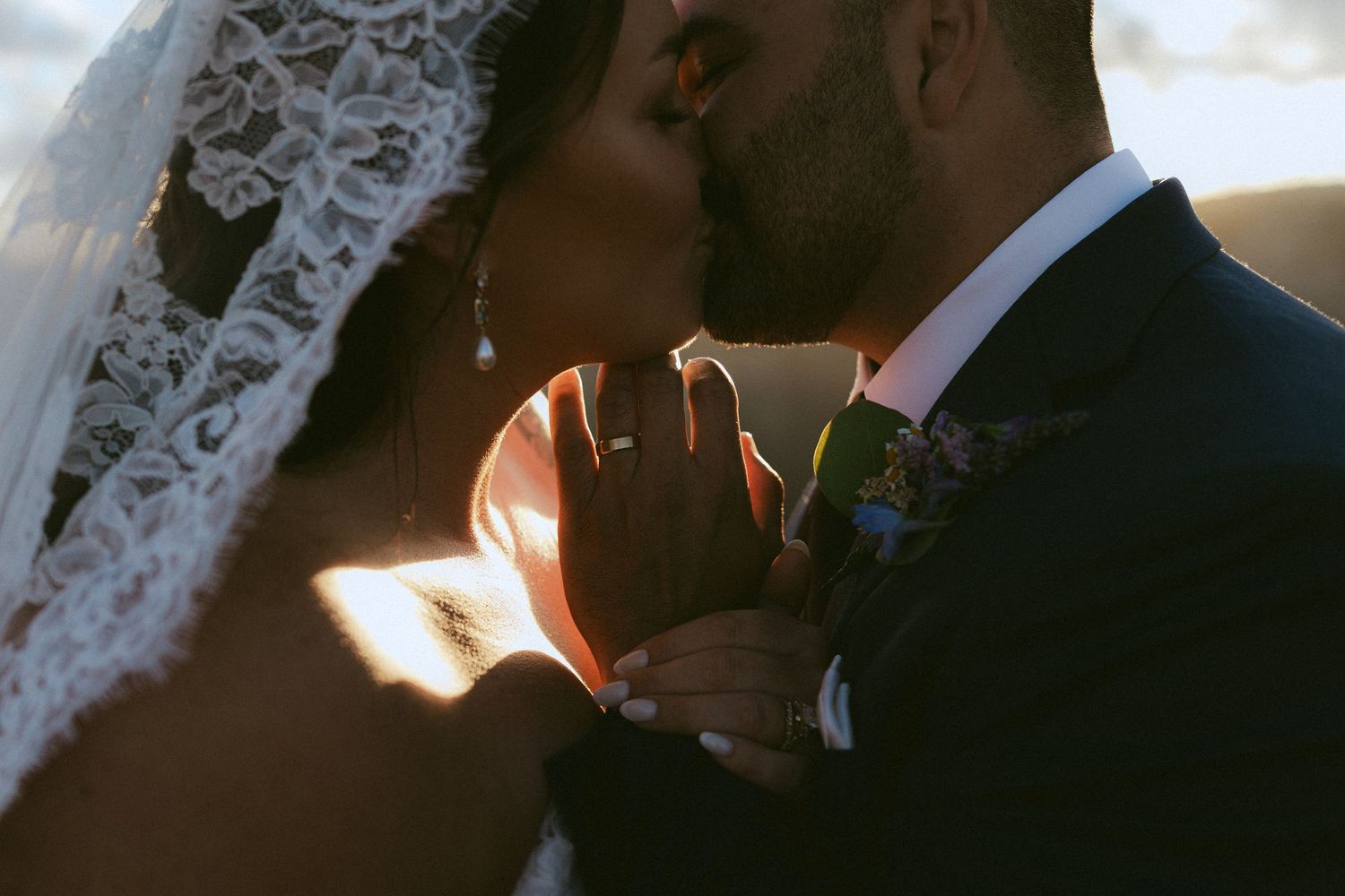 Bride and groom sharing a romantic sunset kiss, bride wearing lace veil, groom in navy suit.