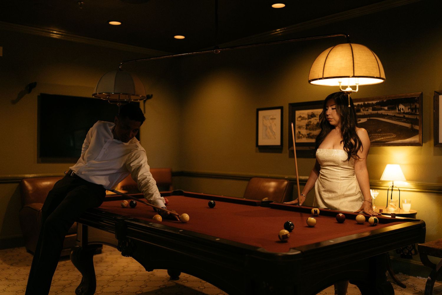 Two people playing pool in a dimly lit game room with a red billiard table under a warm hanging lamp.