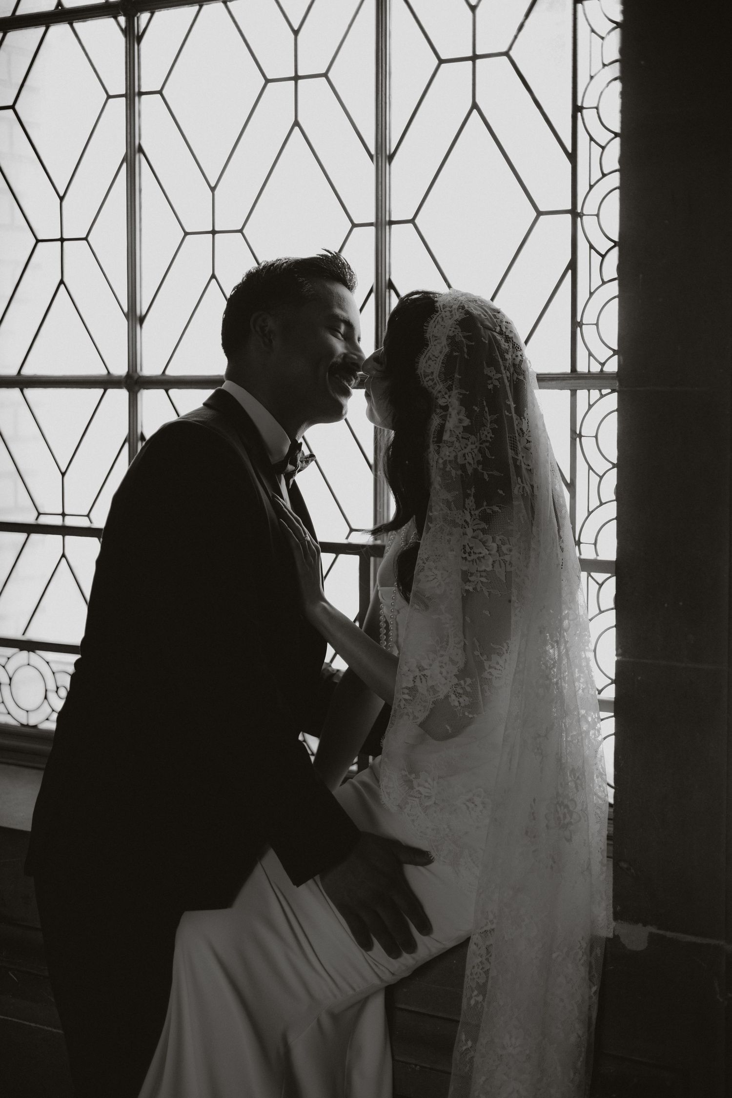 Silhouettes of a bride and groom sharing an intimate moment against a geometric window pattern.