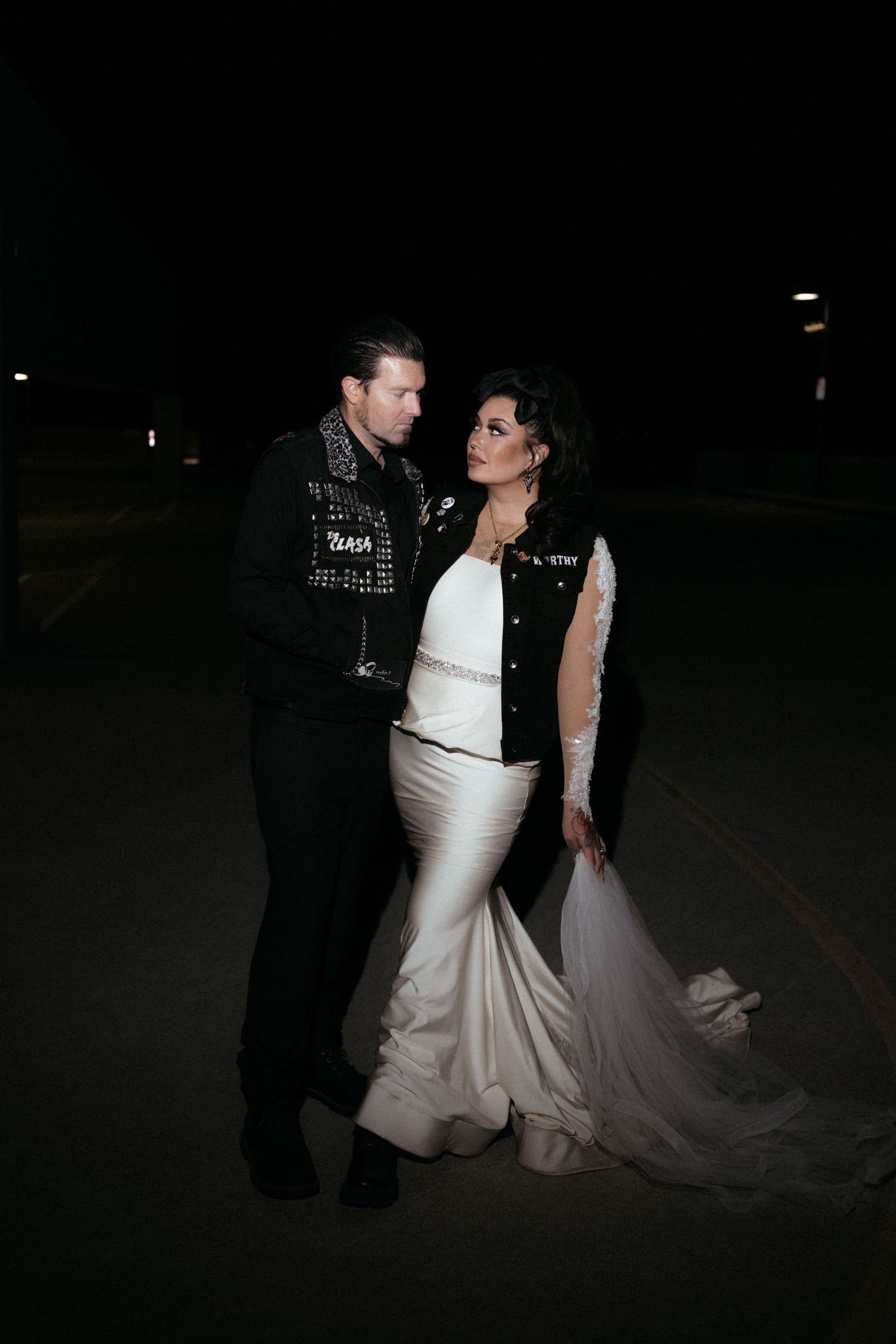 A dramatic nighttime portrait showing the contrast of a white wedding dress against a studded leather jacket.
