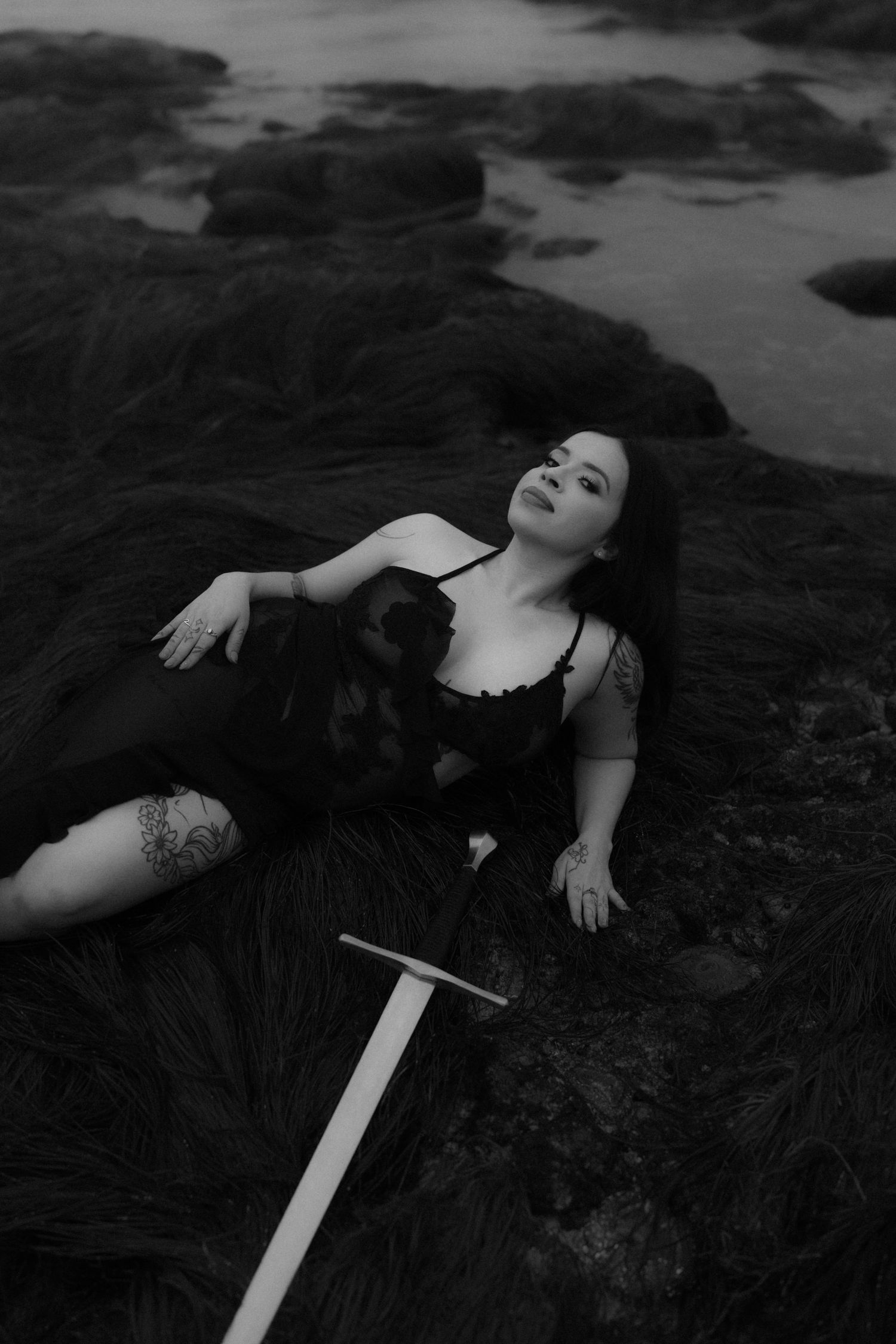 Dark monochrome portrait with medieval sword on rocky beach.