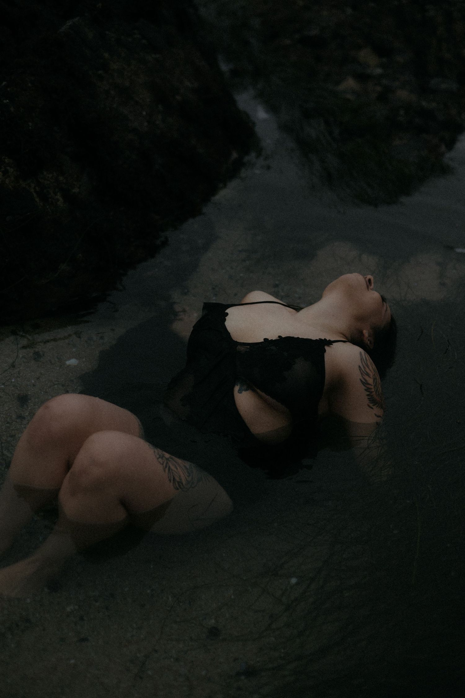Artistic nude photography with figure reclining on dark rocky beach landscape.