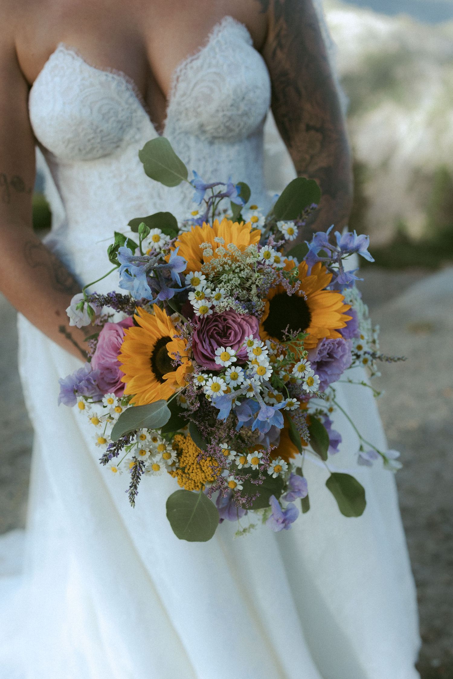 Bride holds vibrant wedding bouquet with sunflowers, purple roses, daisies, and blue wildflowers.